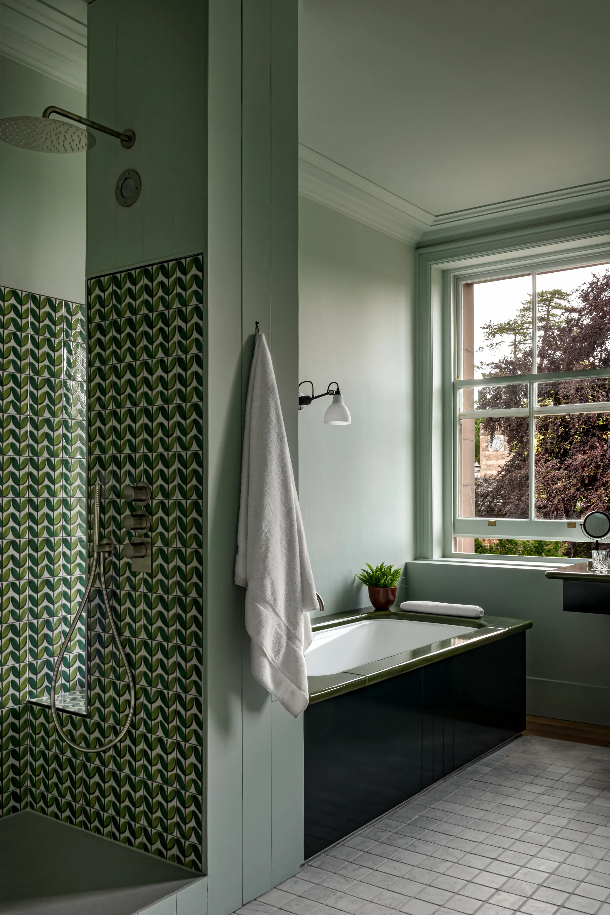 Bathroom with green tiled shower, black-framed bathtub, white towel, potted plant, and large window.