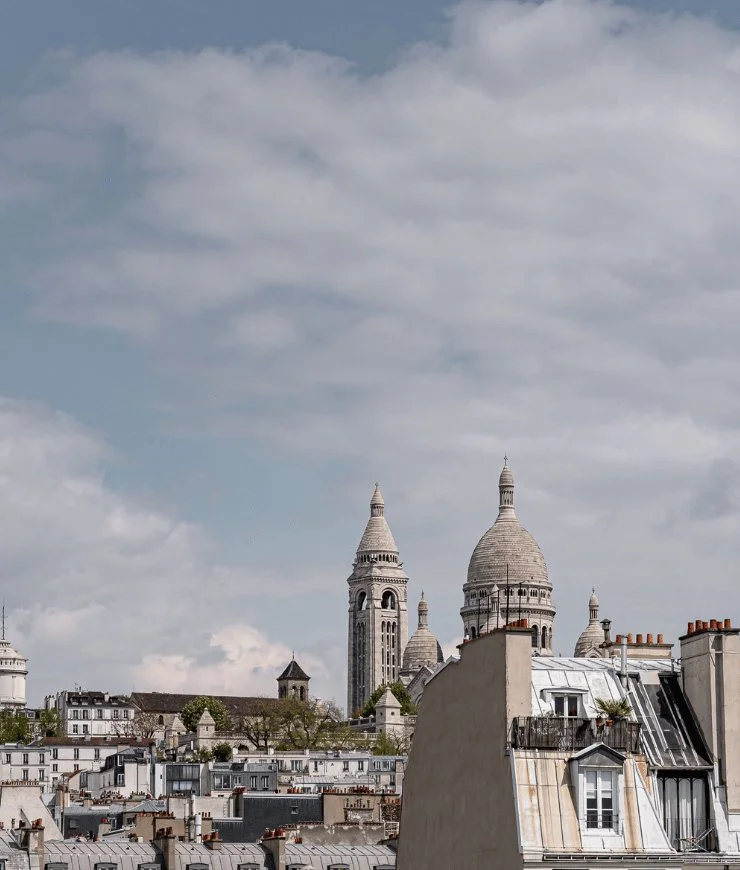 View of Paris rooftops with Sacré-Cœur Basilica domes and towers under cloudy sky.