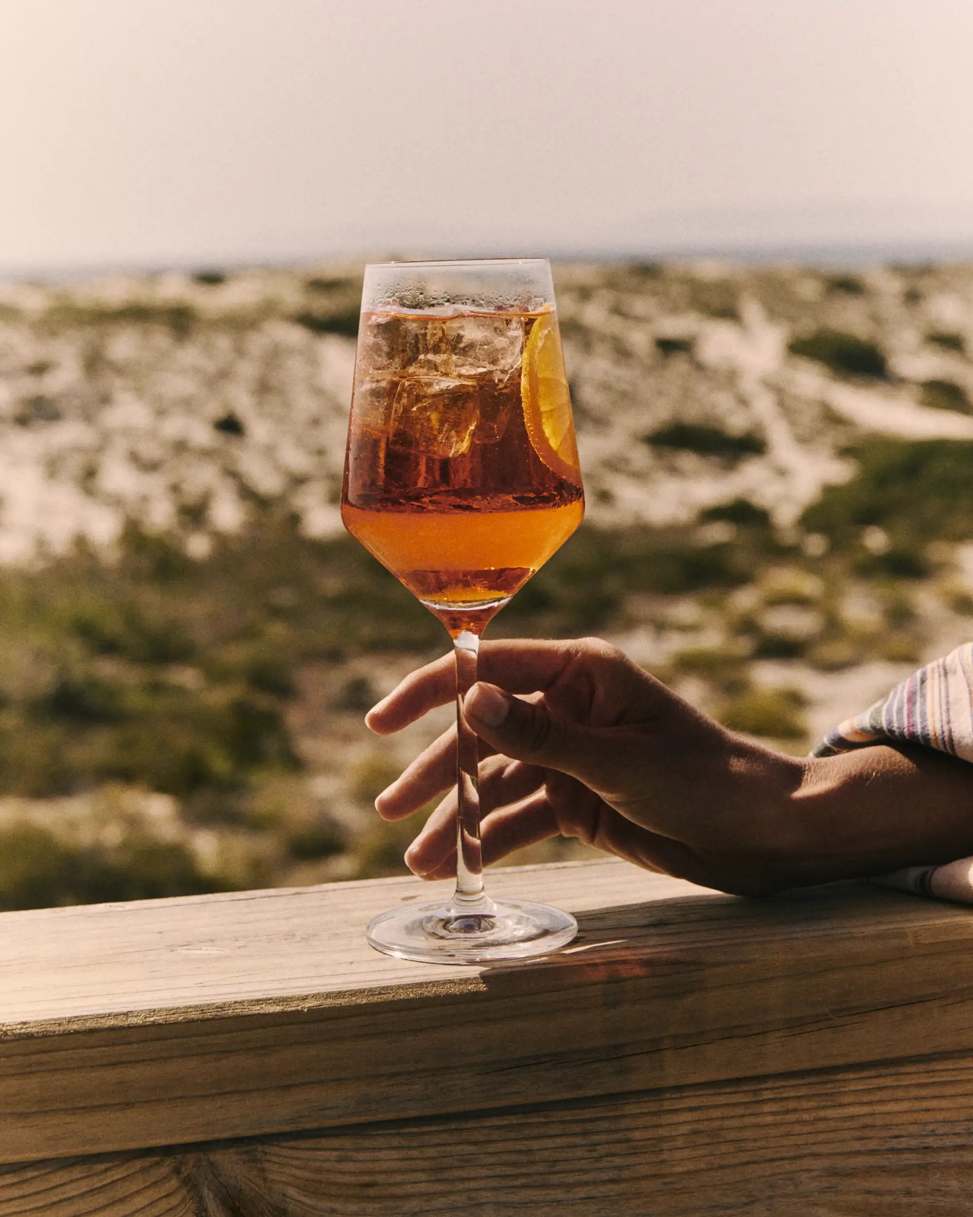 Hand holding a glass of iced orange cocktail with a slice of orange outdoors.