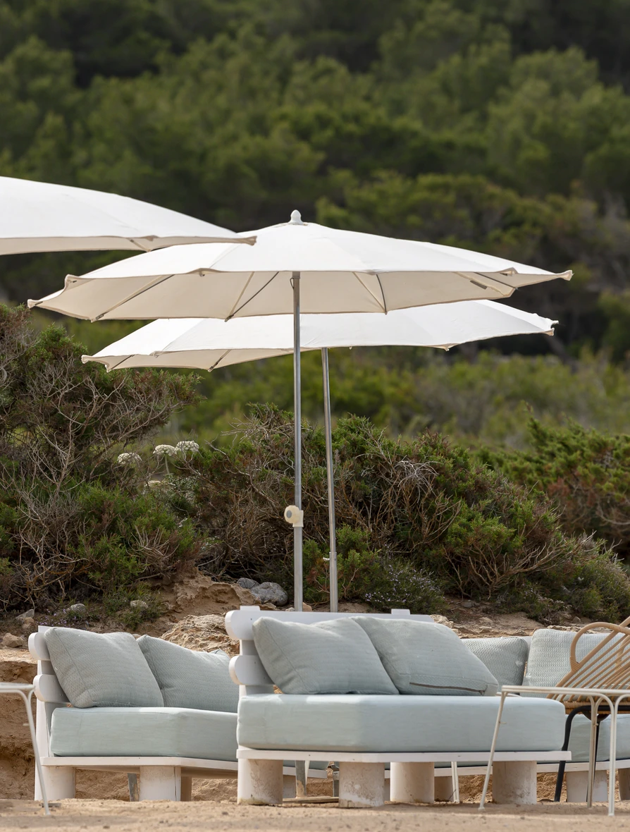 Light-colored outdoor sofas with cushions under white patio umbrellas near green bushes.