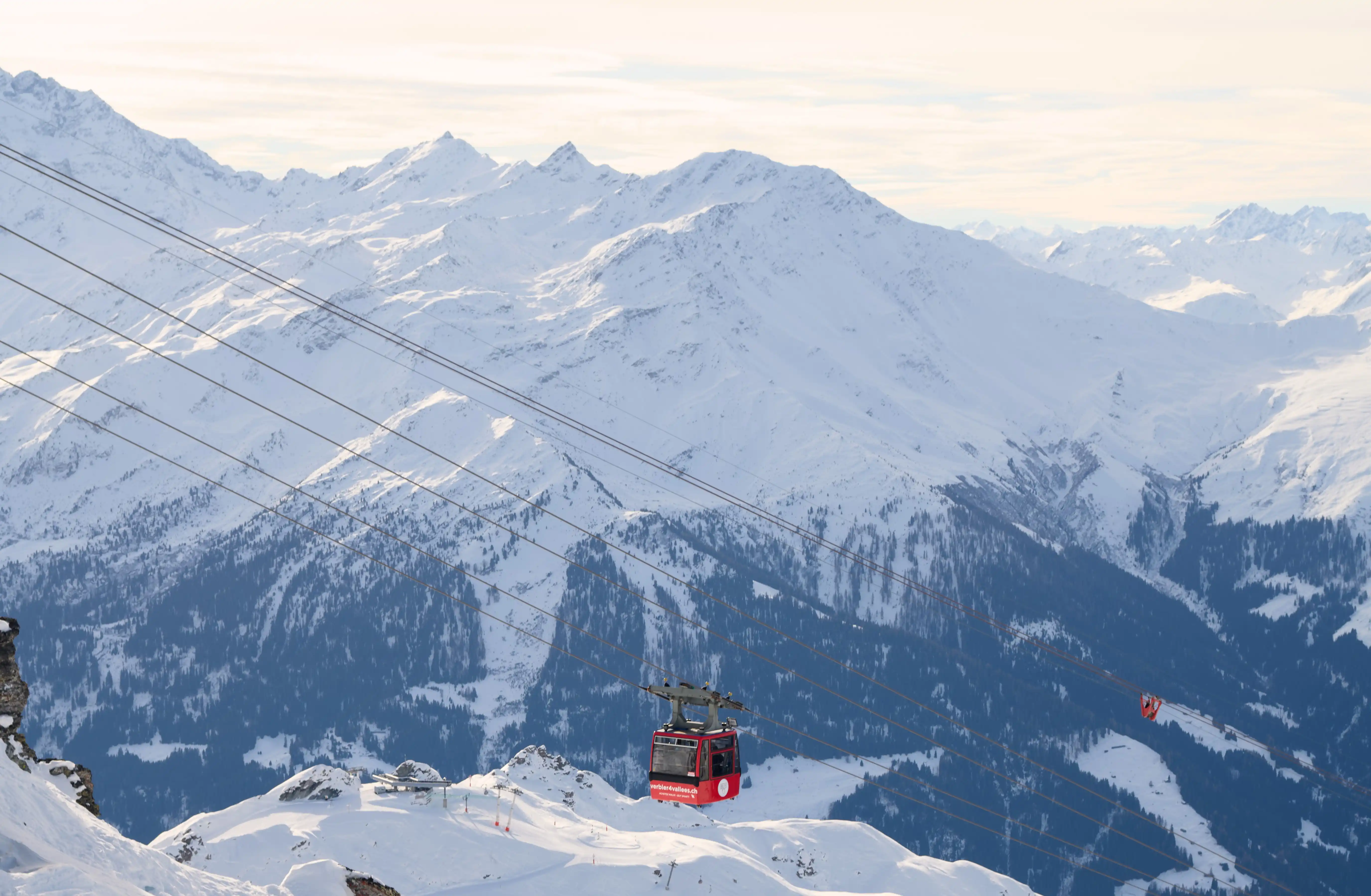 Red cable car on ski lift cables against snowy mountain peaks under a pale sky.