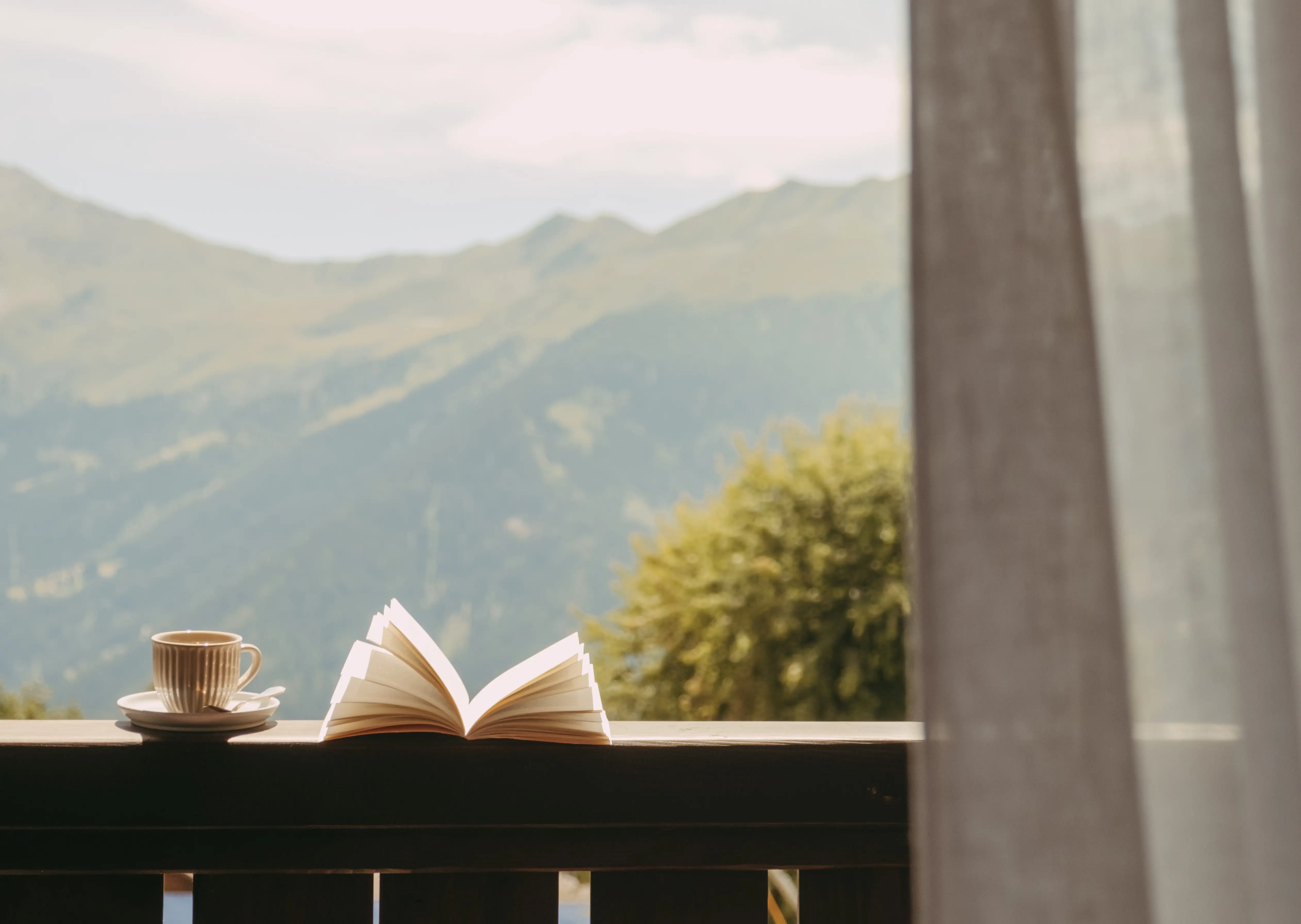 A cup of coffee and an open book on a wooden railing with mountains and trees in the background.