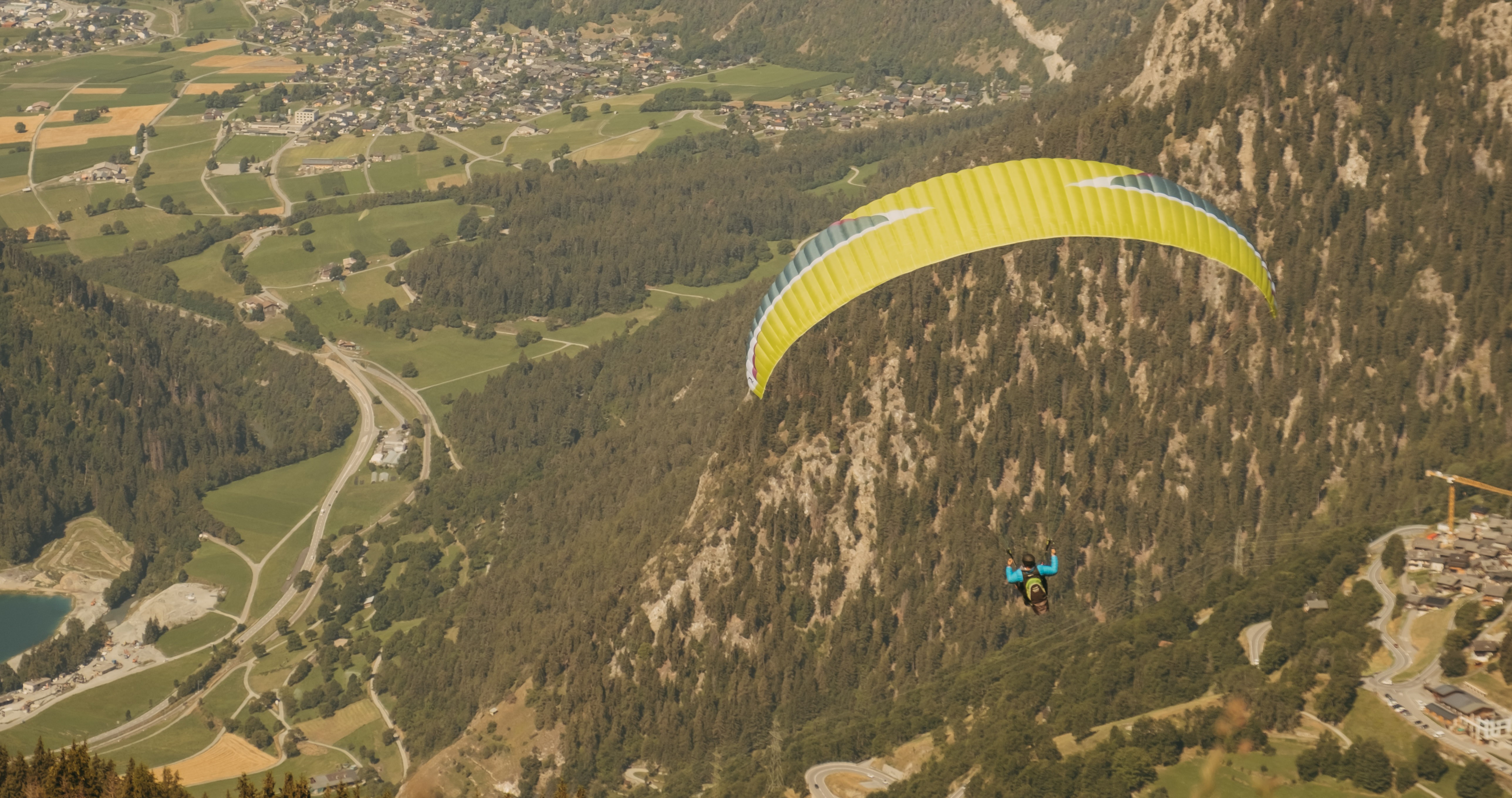 Person paragliding with a yellow parachute near a forested mountain slope and village below.