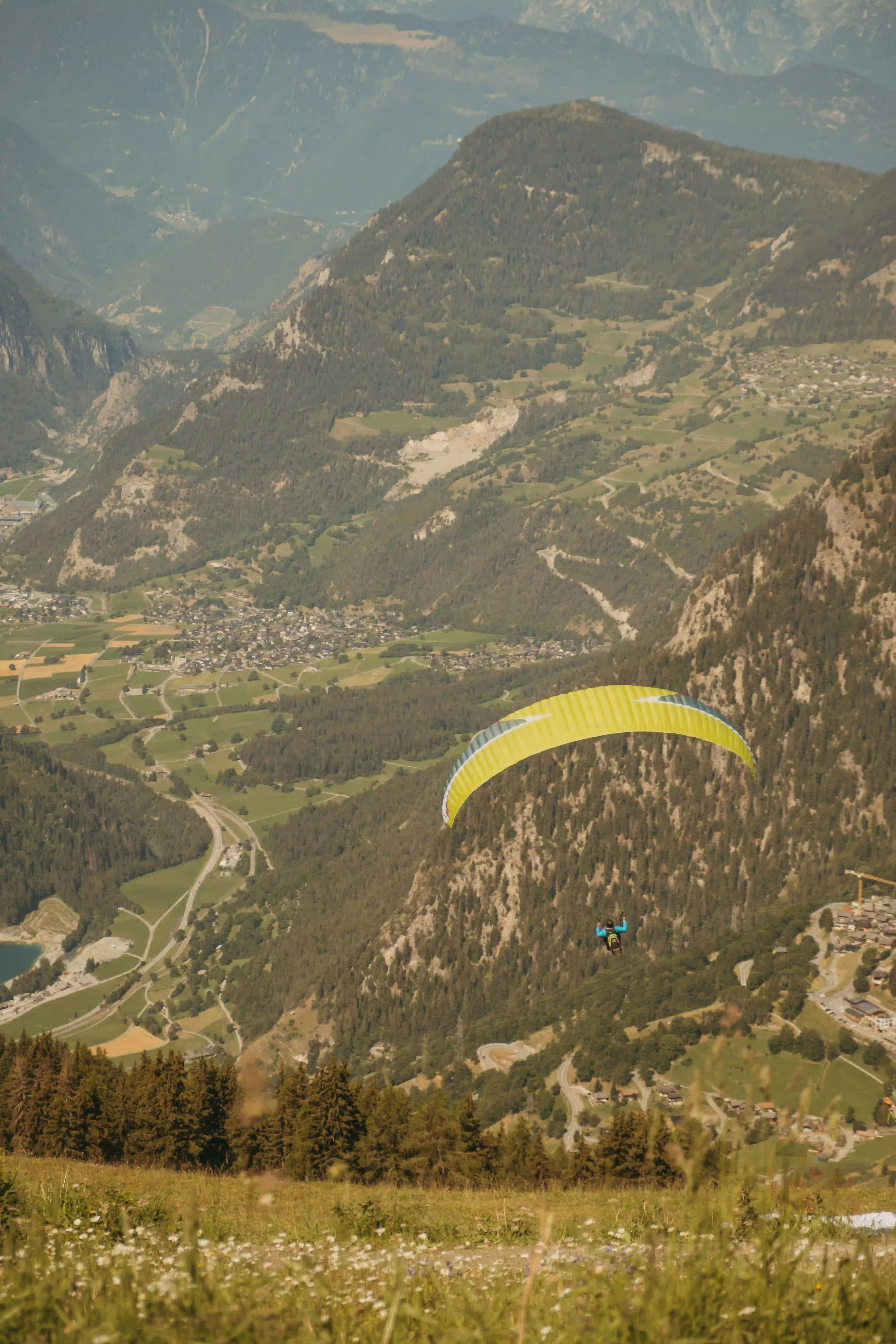 Person paragliding with a yellow parachute over a mountain valley with villages and forests.
