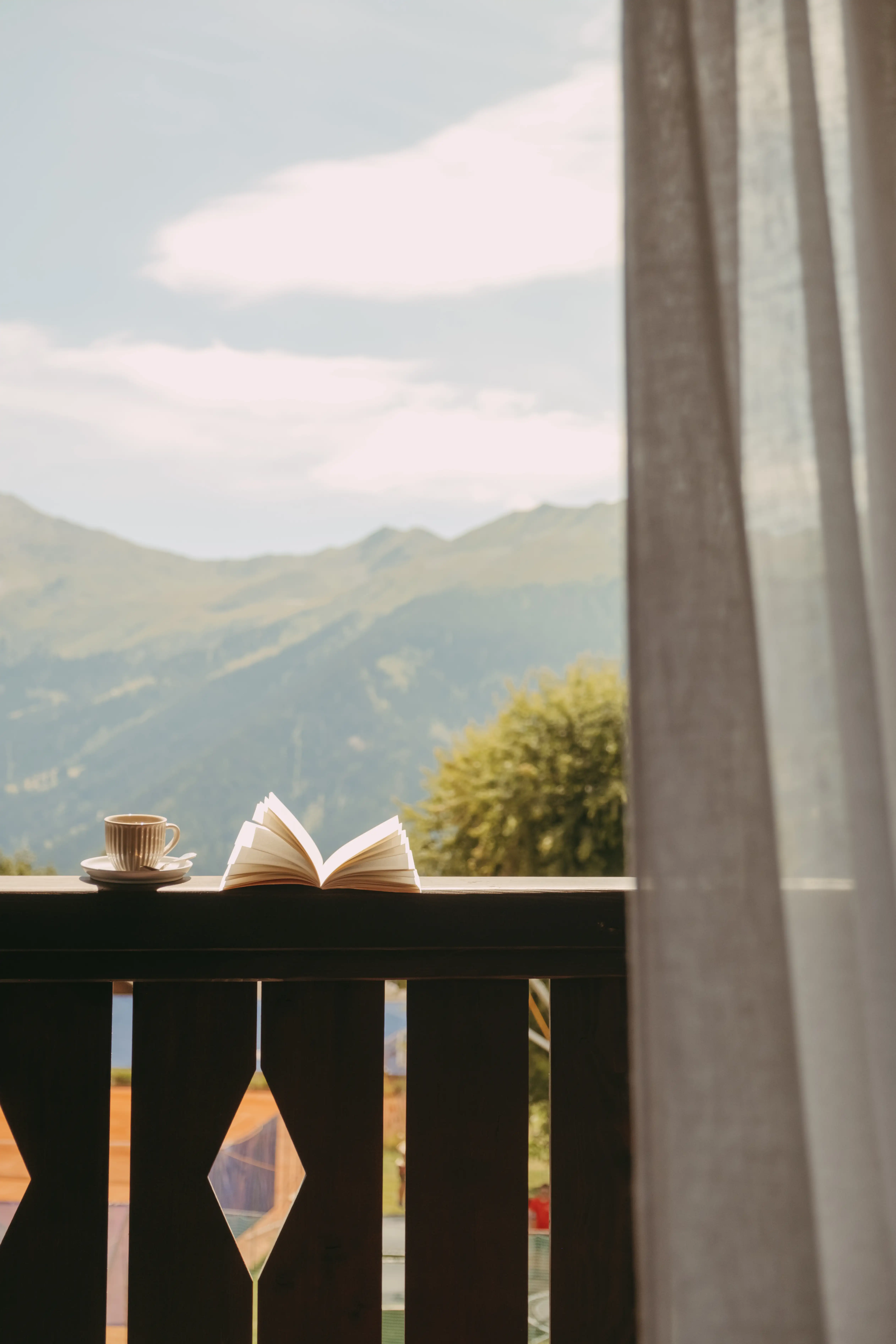 Open book and cup on wooden railing with mountain and tree background behind a sheer curtain.