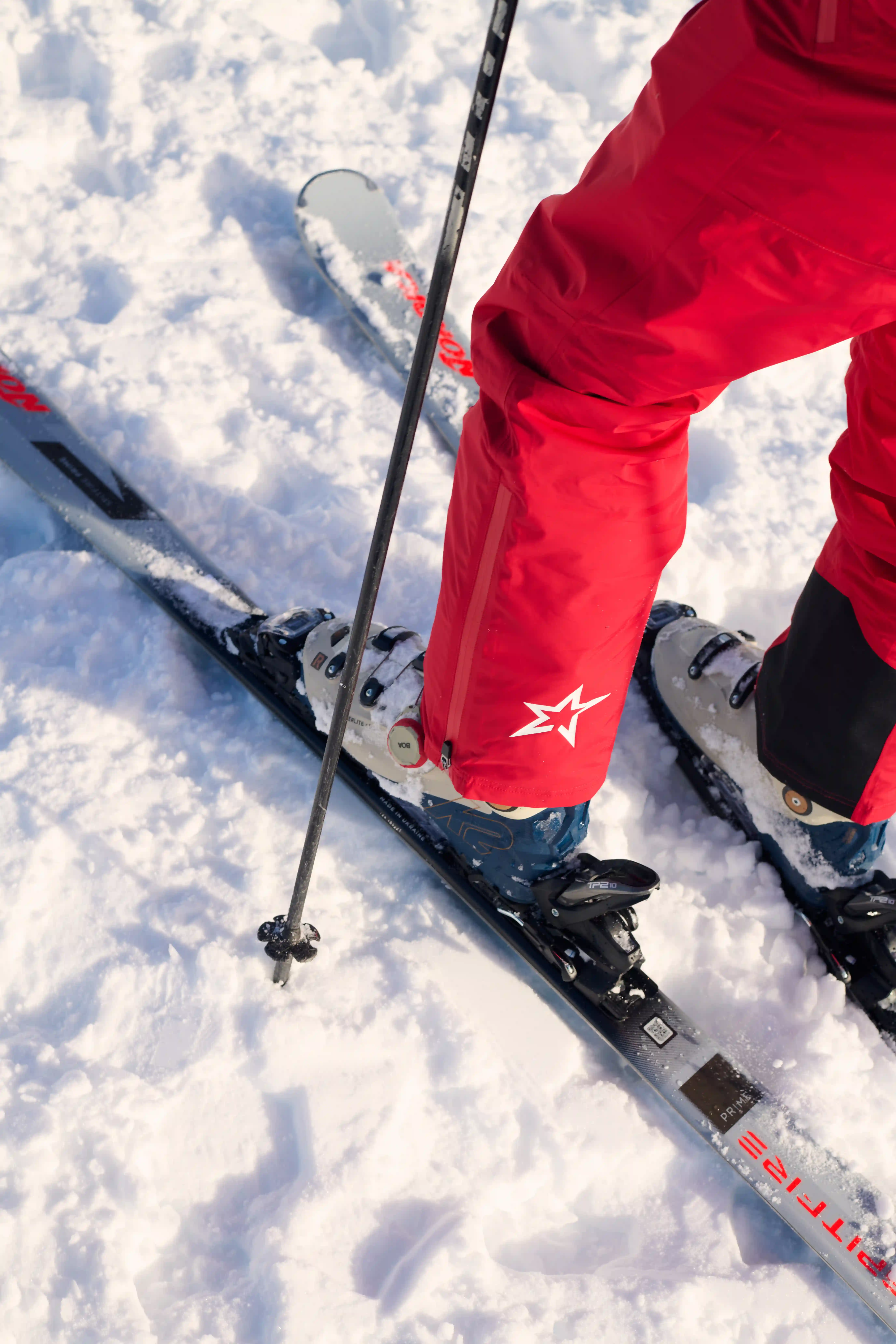 Close-up of skier's legs in red pants and blue boots on skis in snow, holding a ski pole.