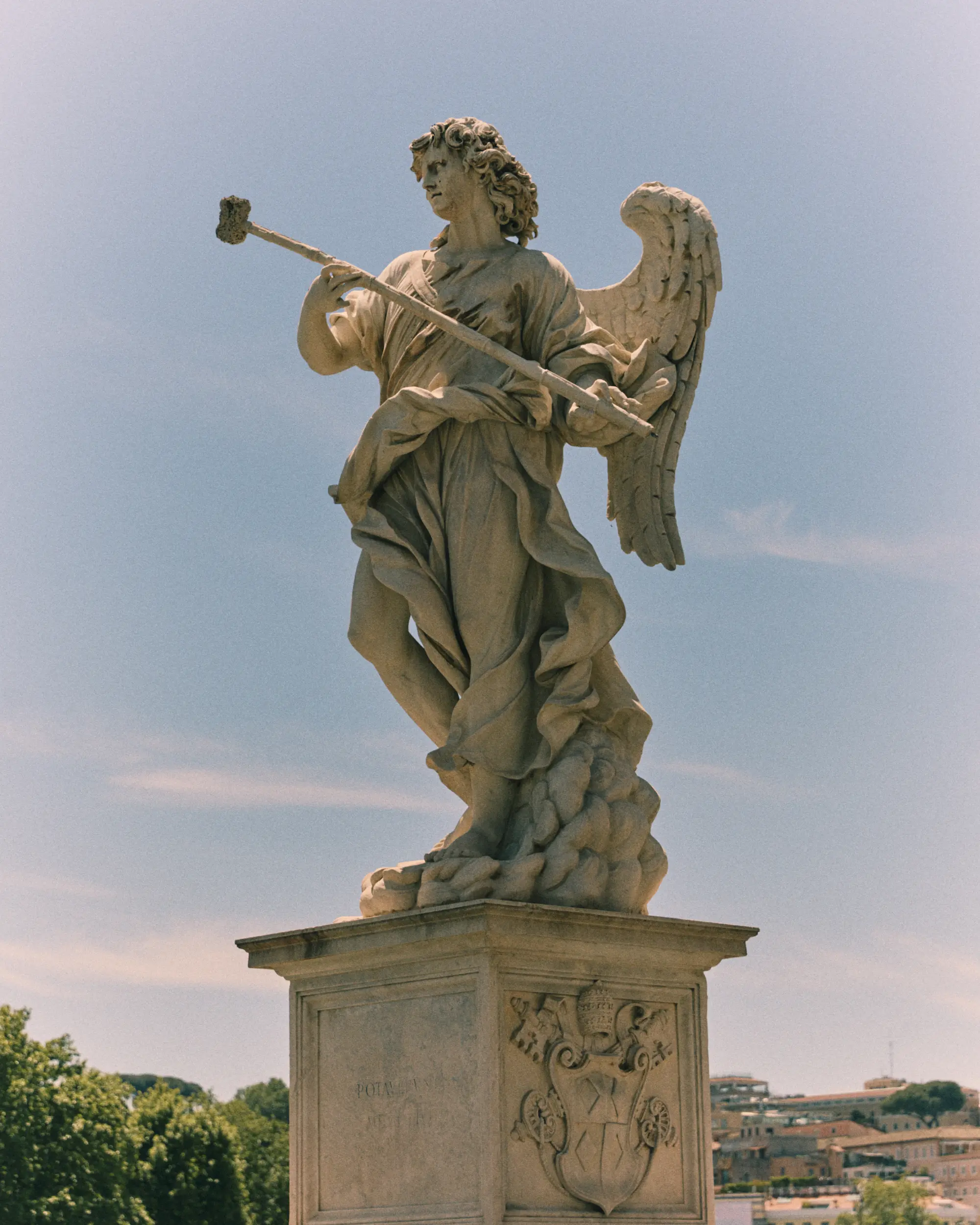 Stone statue of an angel with wings holding a long staff, standing on a pedestal outdoors.