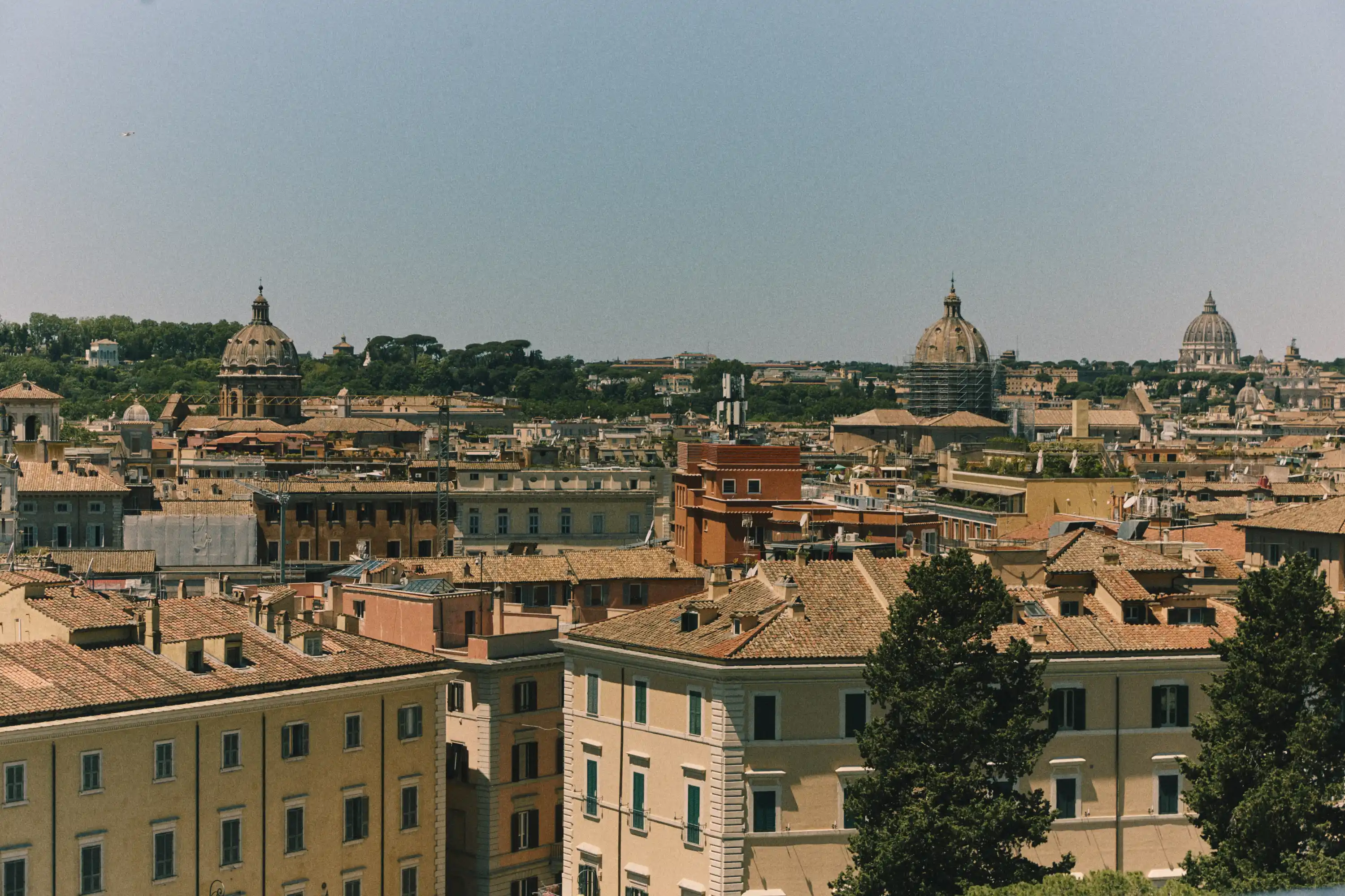 Cityscape of Rome with terracotta rooftops and domed churches under a clear sky.