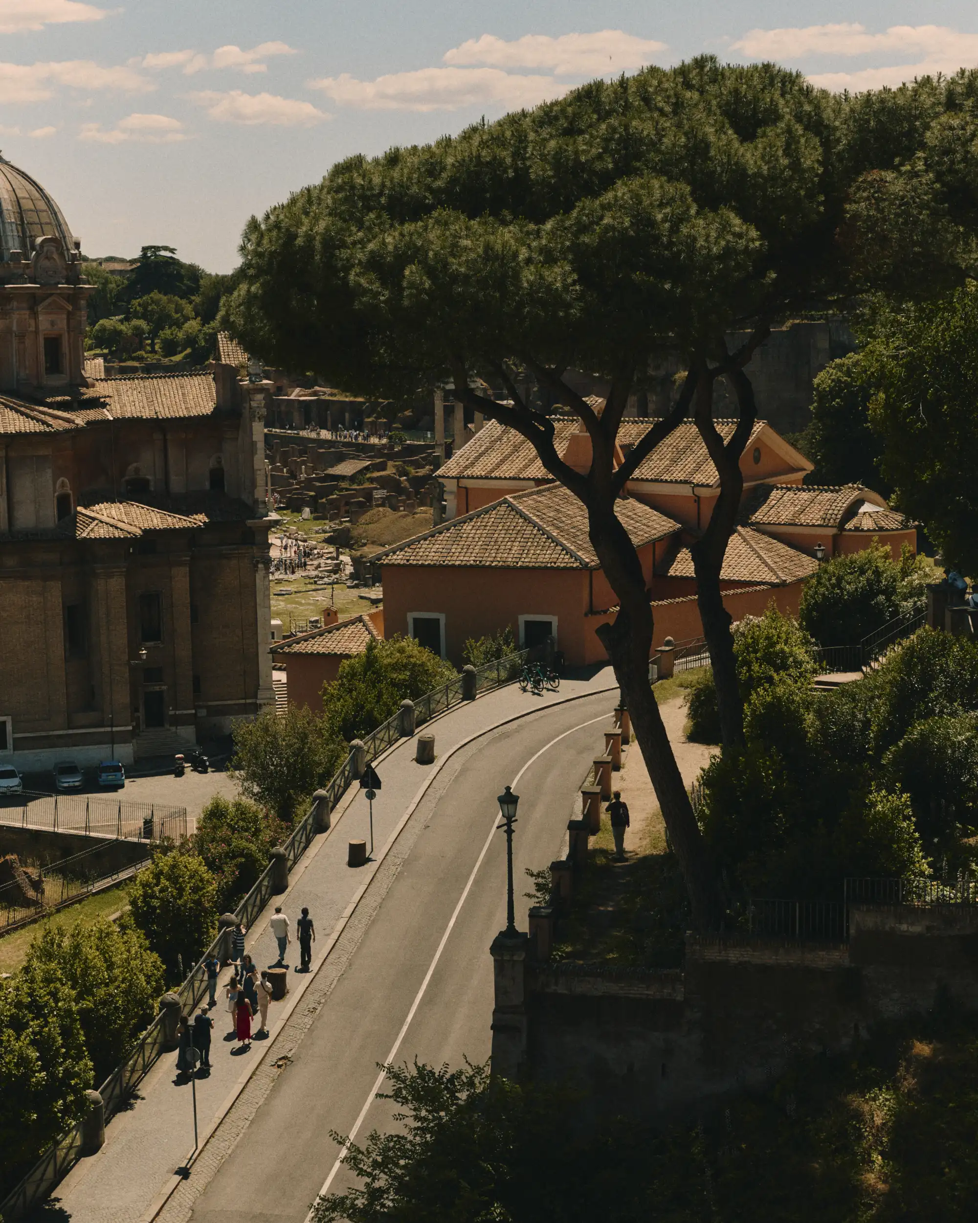 Curved road lined with trees, street lamps, and pedestrians near historic buildings under a partly cloudy sky.