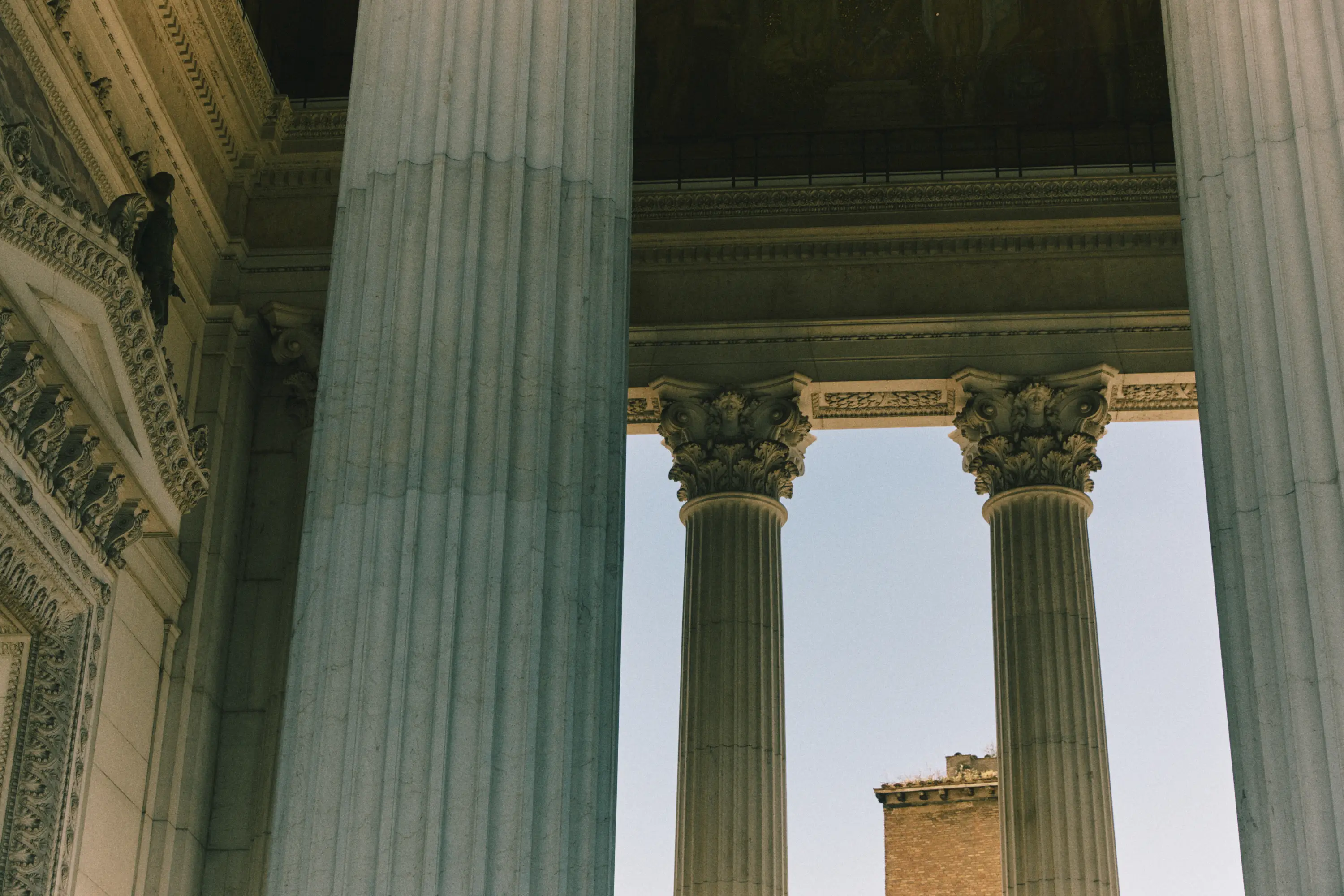 Tall classical columns with ornate Corinthian capitals against a pale sky and partial brick building.