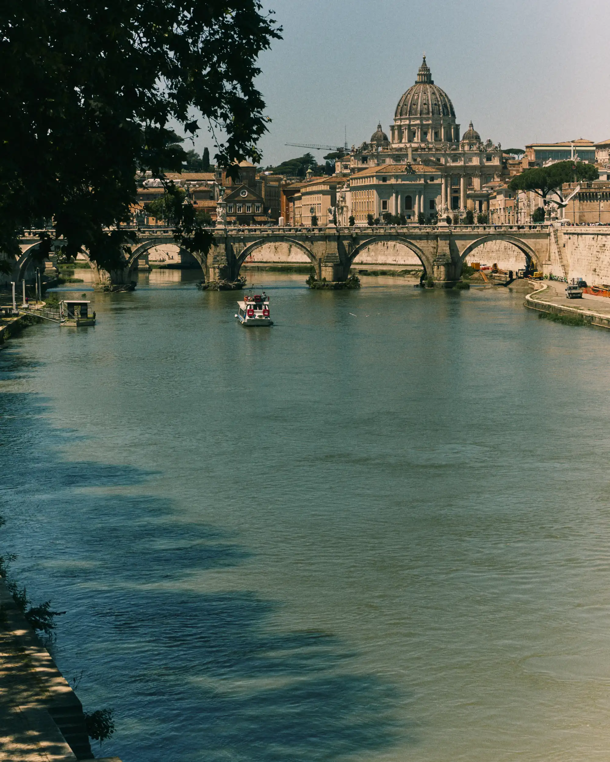 Boat on river with stone bridge and St. Peter's Basilica dome in sunlight in background.