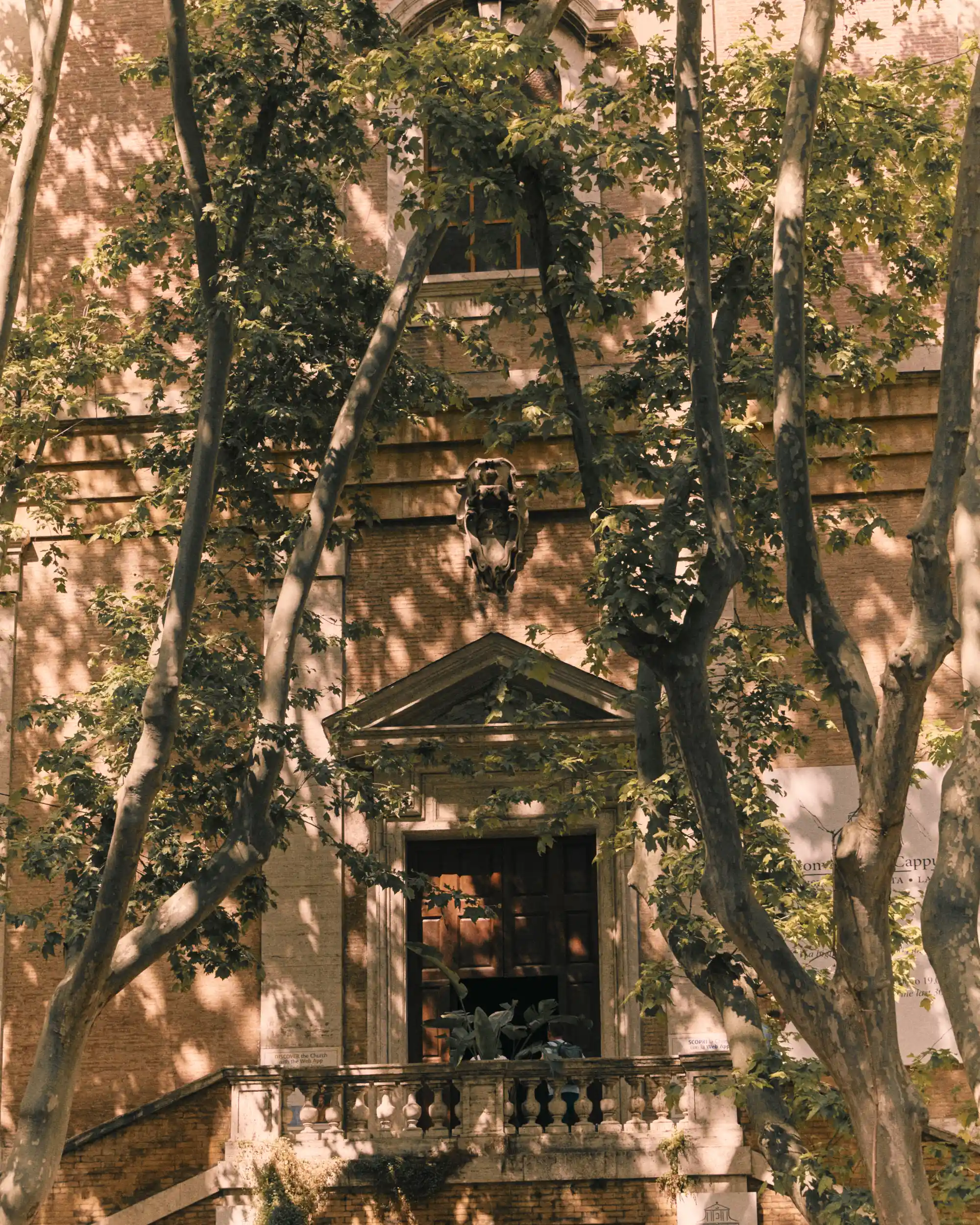 Entrance to a brick building with a balcony, large wooden door, and trees casting shadows.