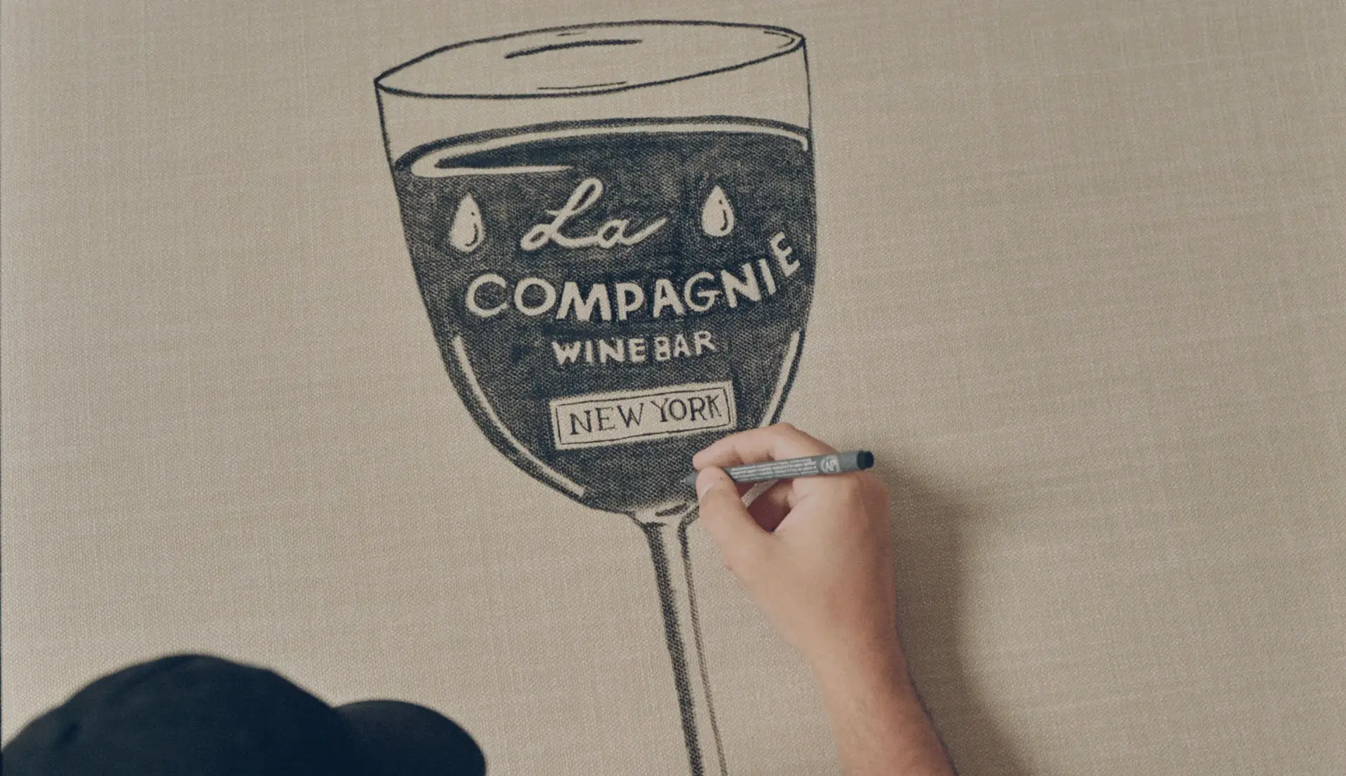 Desenho manual de uma taça de vinho com o texto “La Compagnie Wine Bar New York” em uma tela bege.