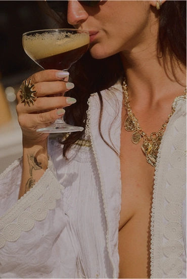 Mulher de túnica branca tomando uma bebida escura e espumosa em um copo de coquetel, usando joias de ouro e anel.
