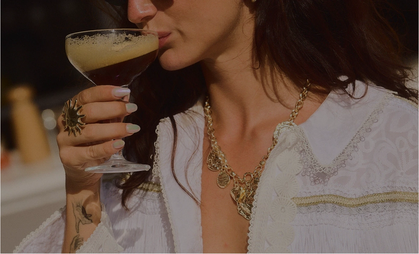 Mulher de blusa branca com colar de ouro bebendo uma bebida espumosa escura em um copo de coquetel.