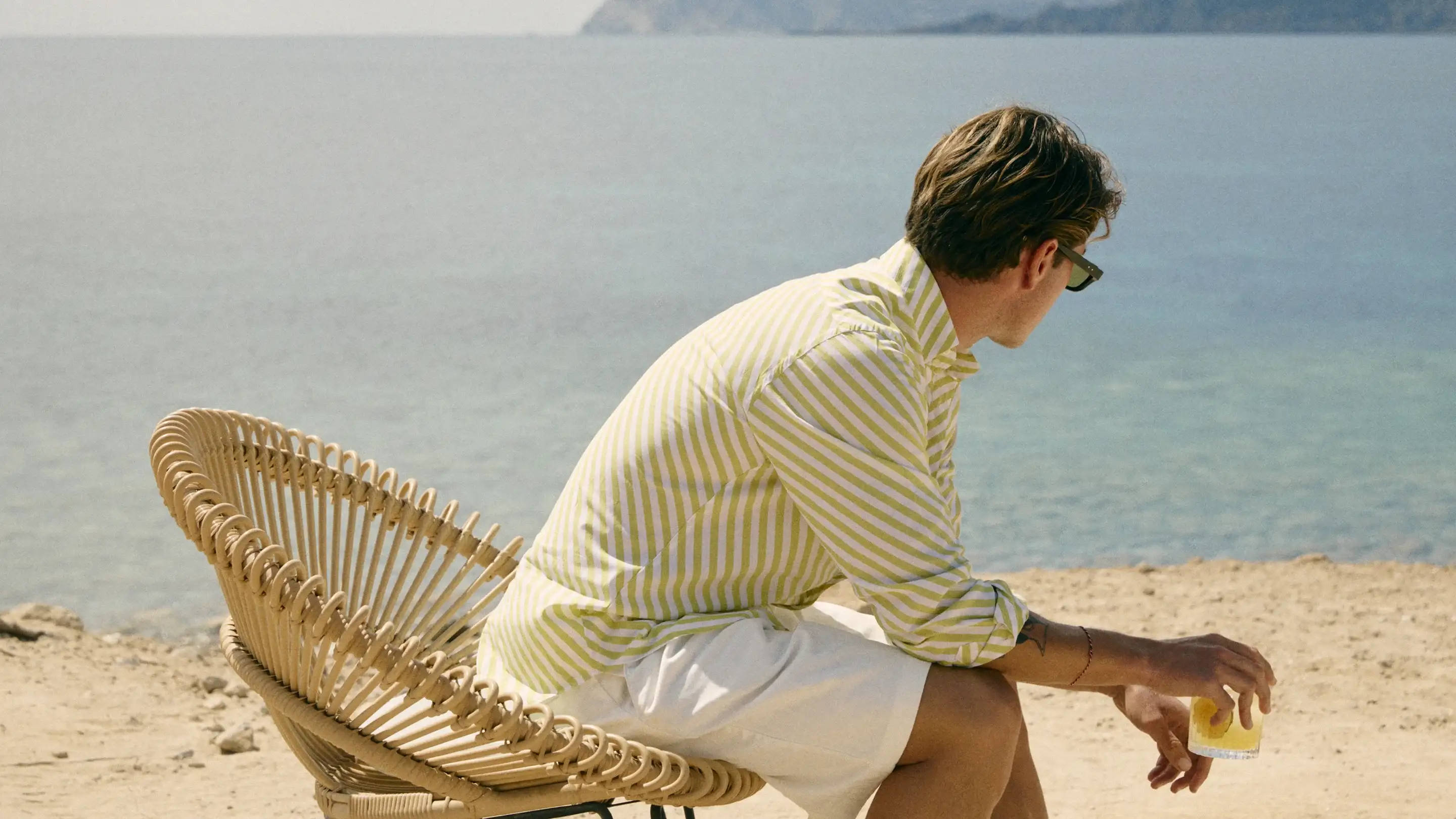 Man in striped shirt and white shorts sitting on beach chair, holding a glass, facing the sea.