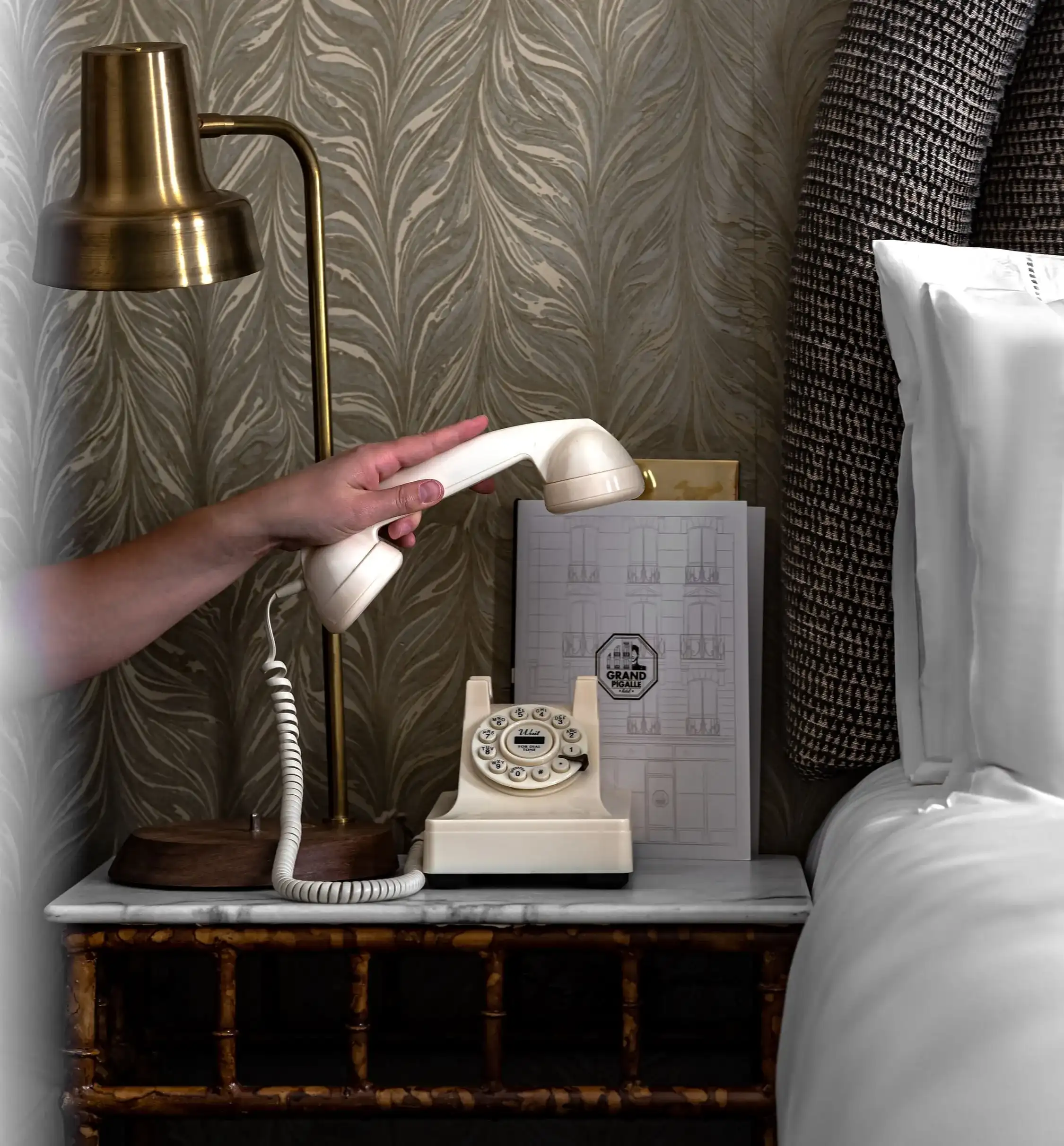 Hand lifting handset of vintage cream rotary dial phone on bedside table next to lamp and bed.