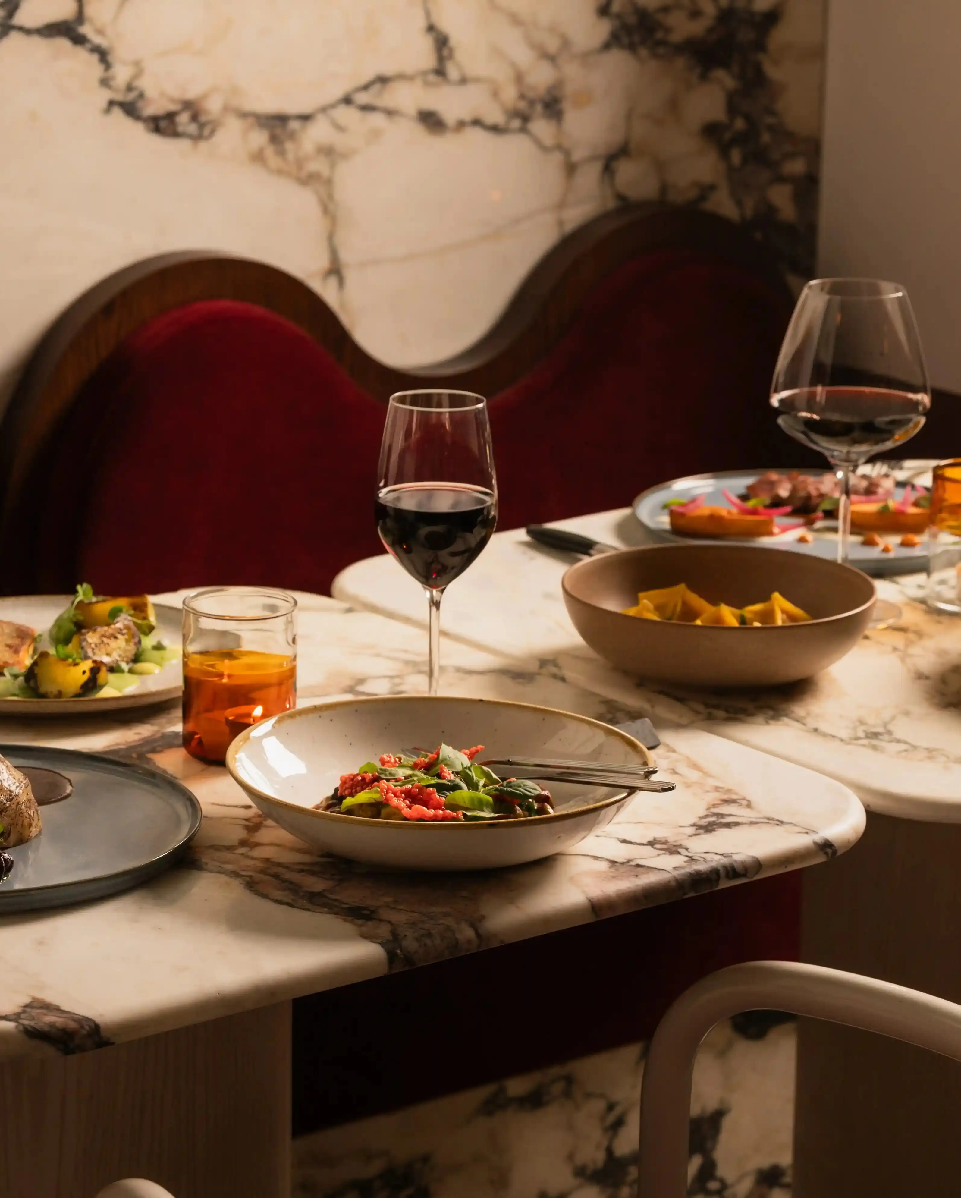 Mesa de mármore com pratos gourmet revestidos, taças de vinho com vinho tinto e taças de água de cor âmbar.