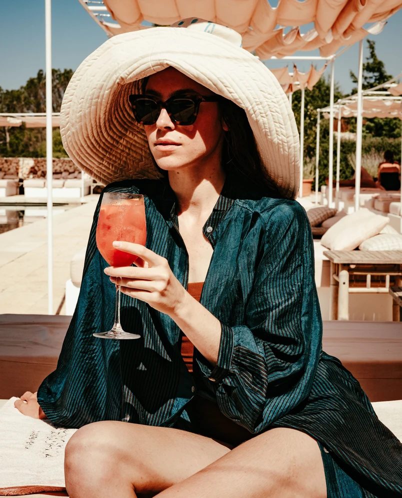 Woman in a large sun hat and sunglasses holding a glass of orange cocktail by a sunny poolside lounge.