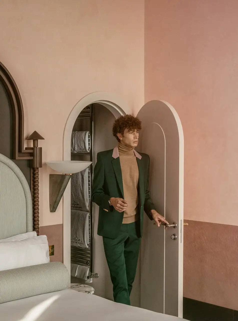 Man in a green suit and brown turtleneck walking through a rounded doorway in a pastel room.