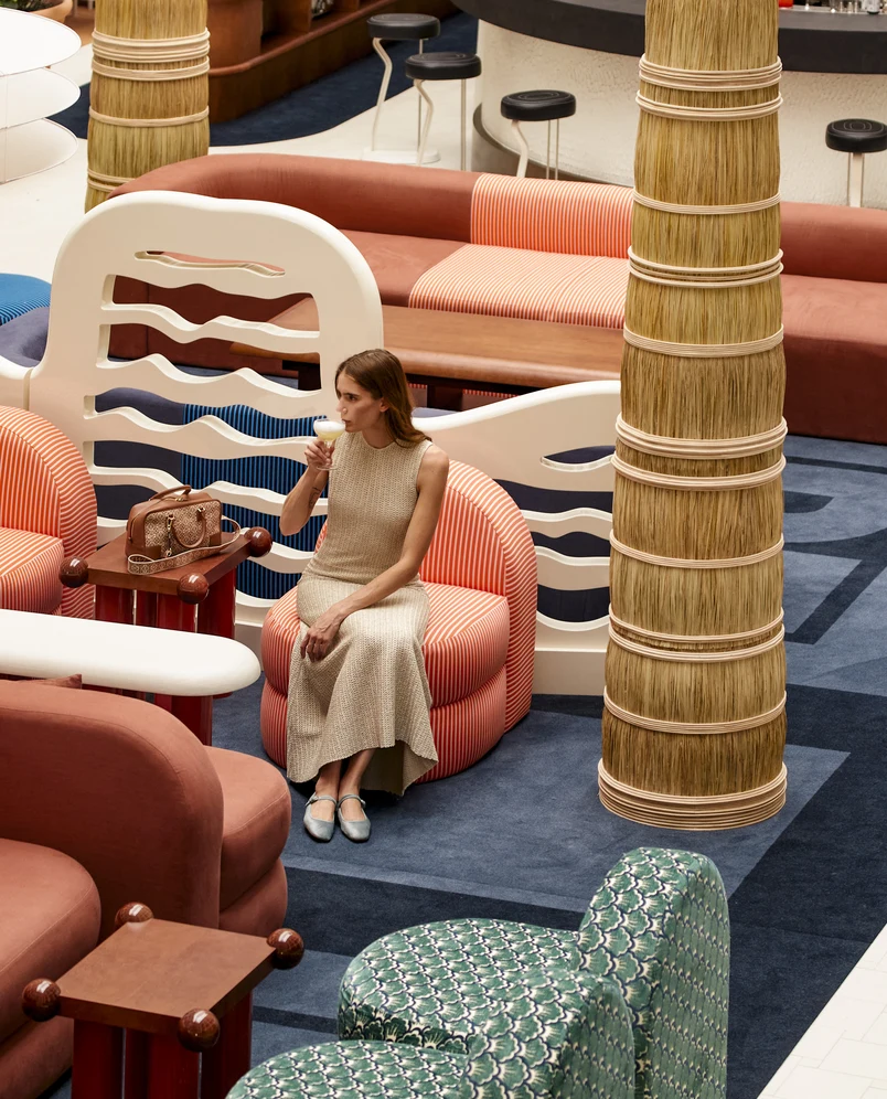 Mujer vestida de color beige sentada en una silla a rayas bebiendo un cóctel en un colorido y moderno salón.