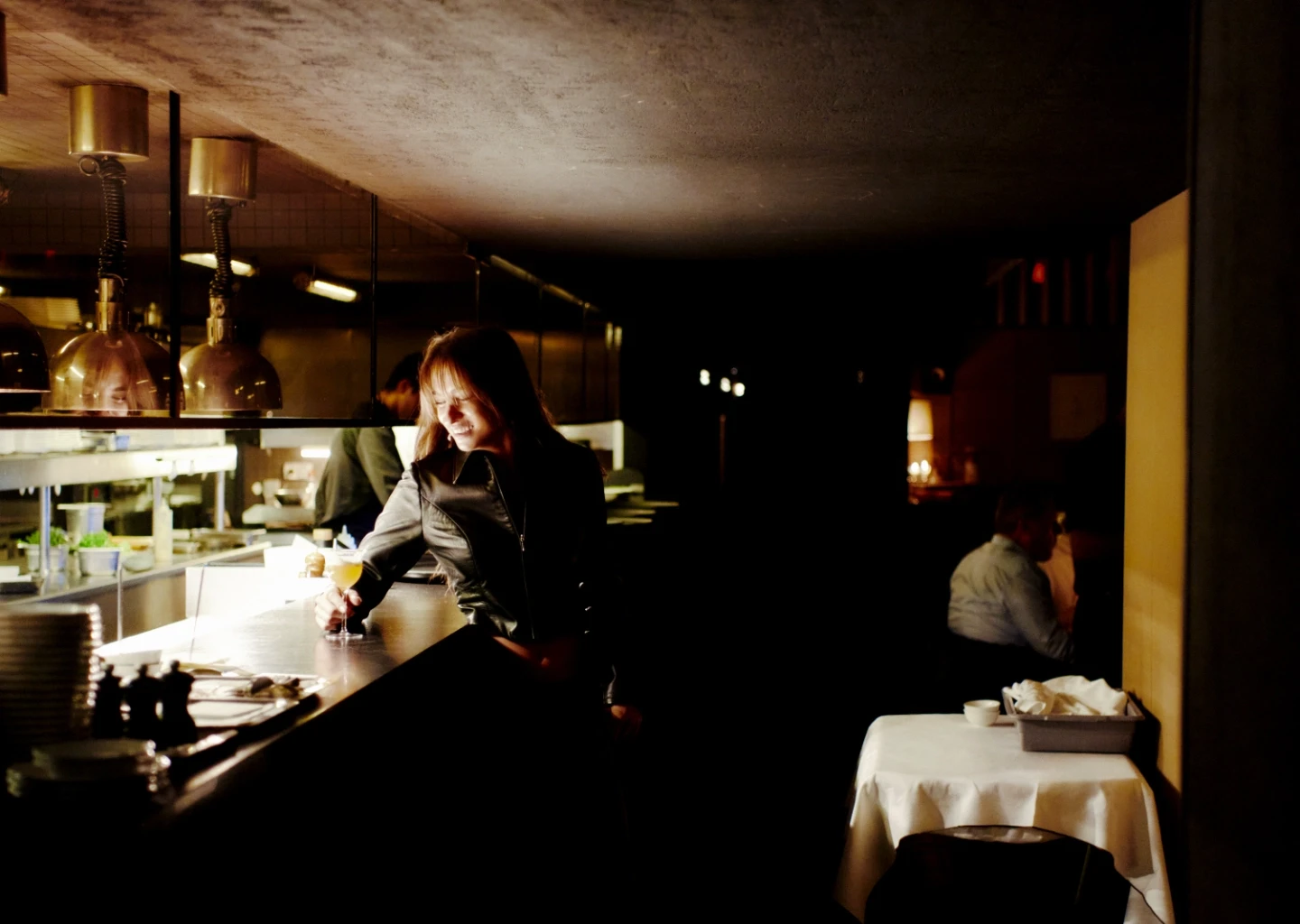 Donna in una giacca di pelle sorridente mentre tiene in mano un drink in un bar poco illuminato con cucina sullo sfondo.