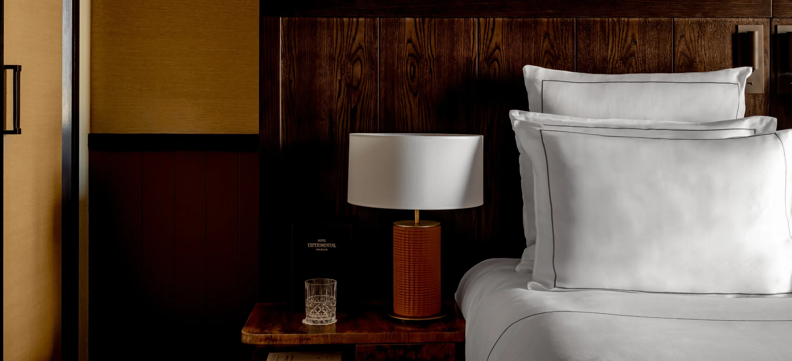 Comodino con lampada, vetro e menu dell'hotel accanto a cuscini bianchi sul letto con testiera in legno