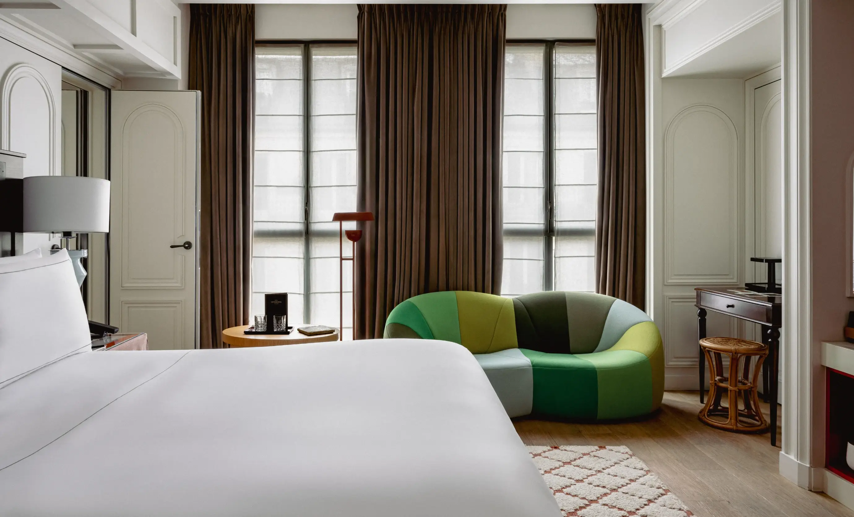 Camera d'albergo con letto bianco, divano verde multicolore, pavimento in legno, tavolino e ampie finestre con tende marroni.