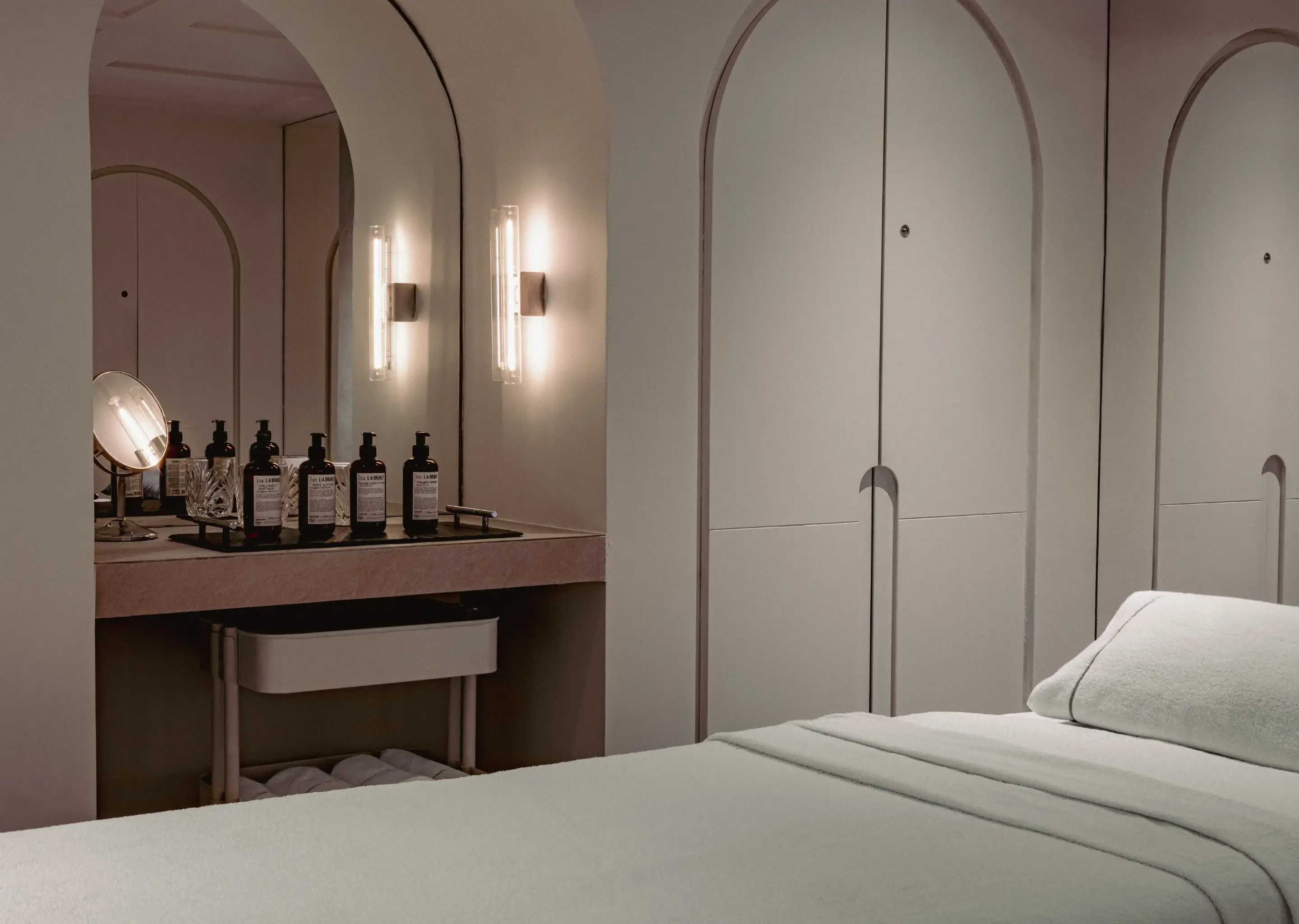 Tranquilla sala per trattamenti spa con lettino da massaggio, specchio ad arco, lampade e articoli da toeletta su un bancone.