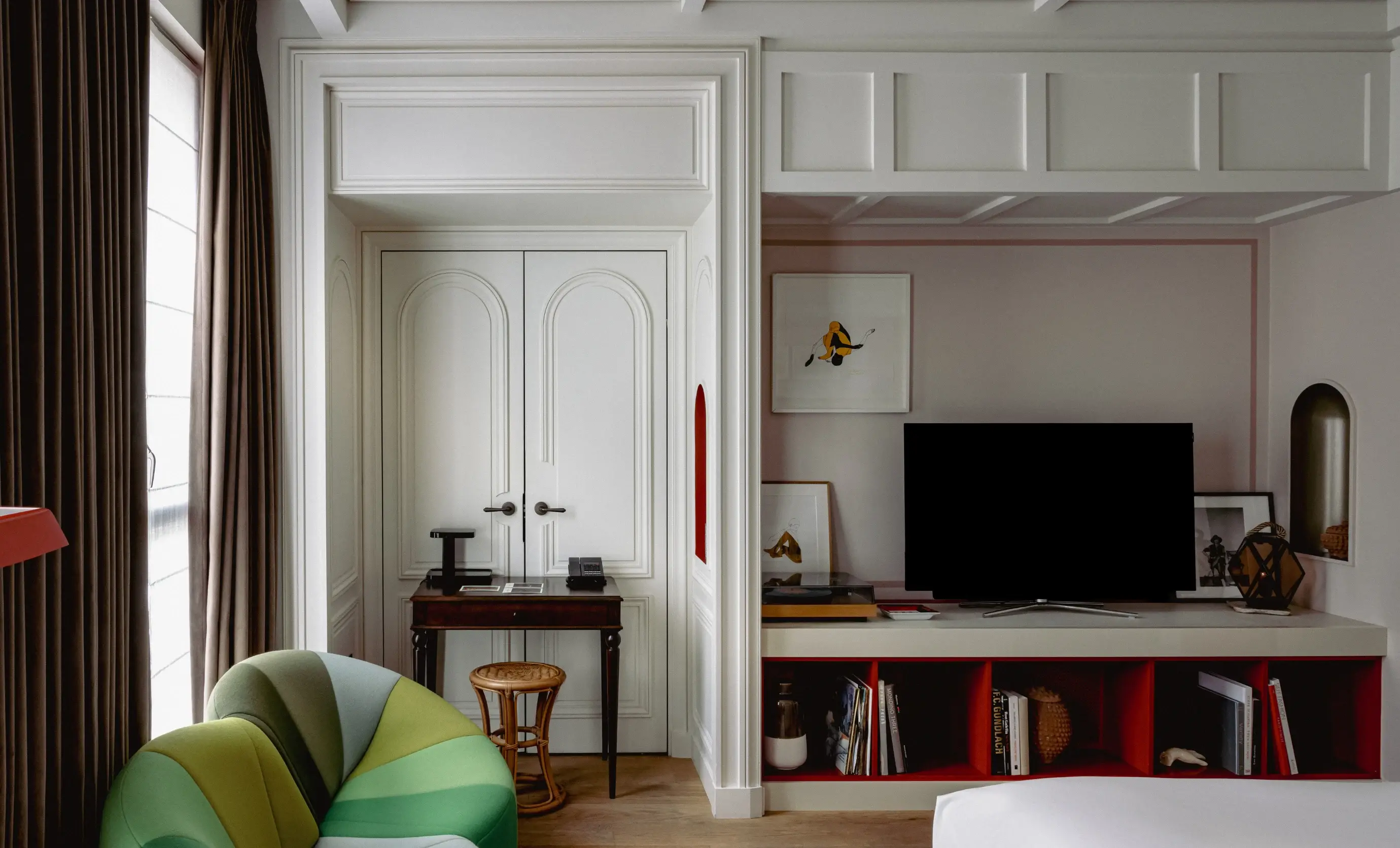 Camera d'albergo con sedia curva verde, doppie porte bianche, scrivania, TV su scaffale con libri e decorazioni.