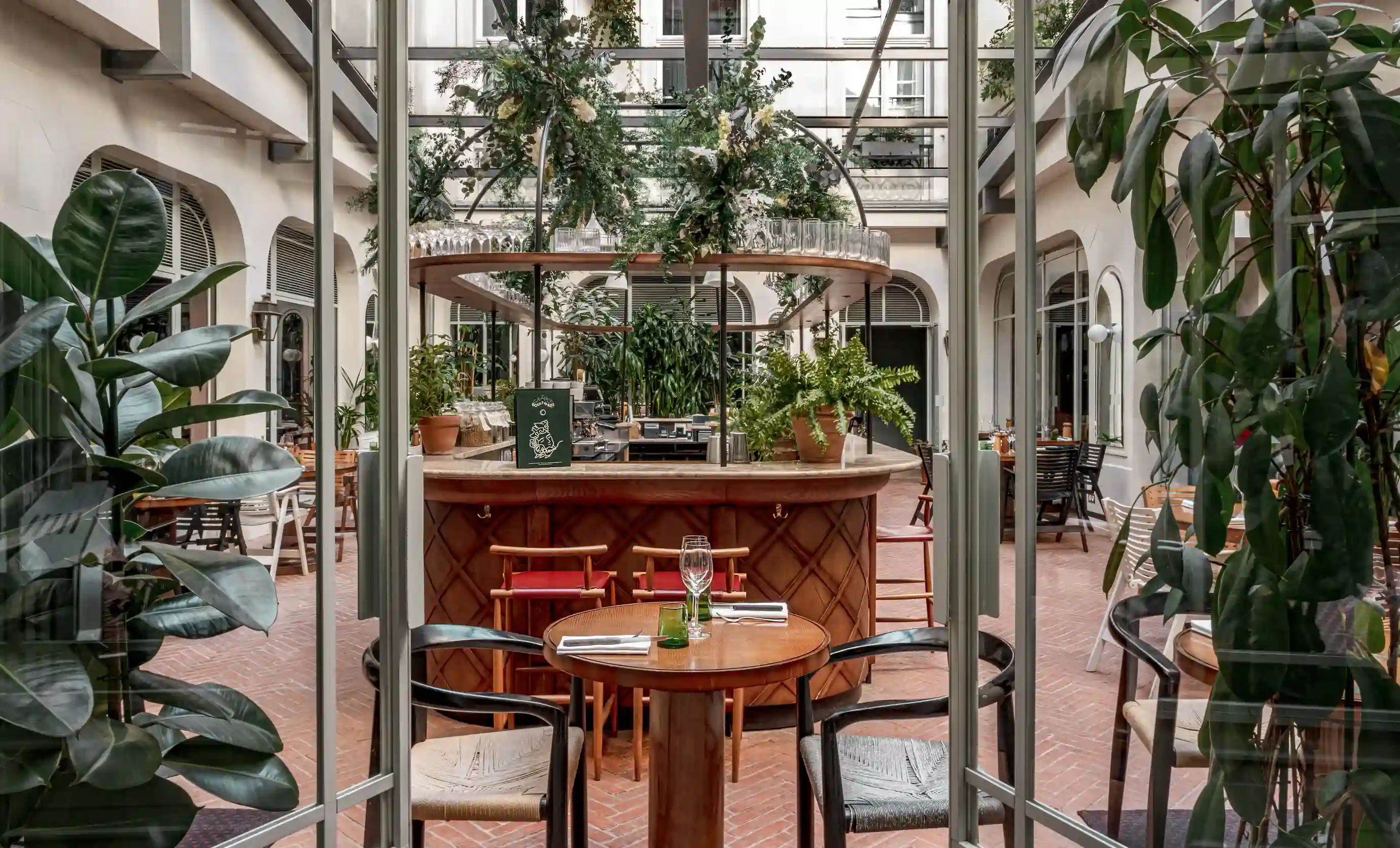 Interior aconchegante do restaurante com mesas de madeira, cadeiras e um bar central decorado com plantas sob um teto de vidro.
