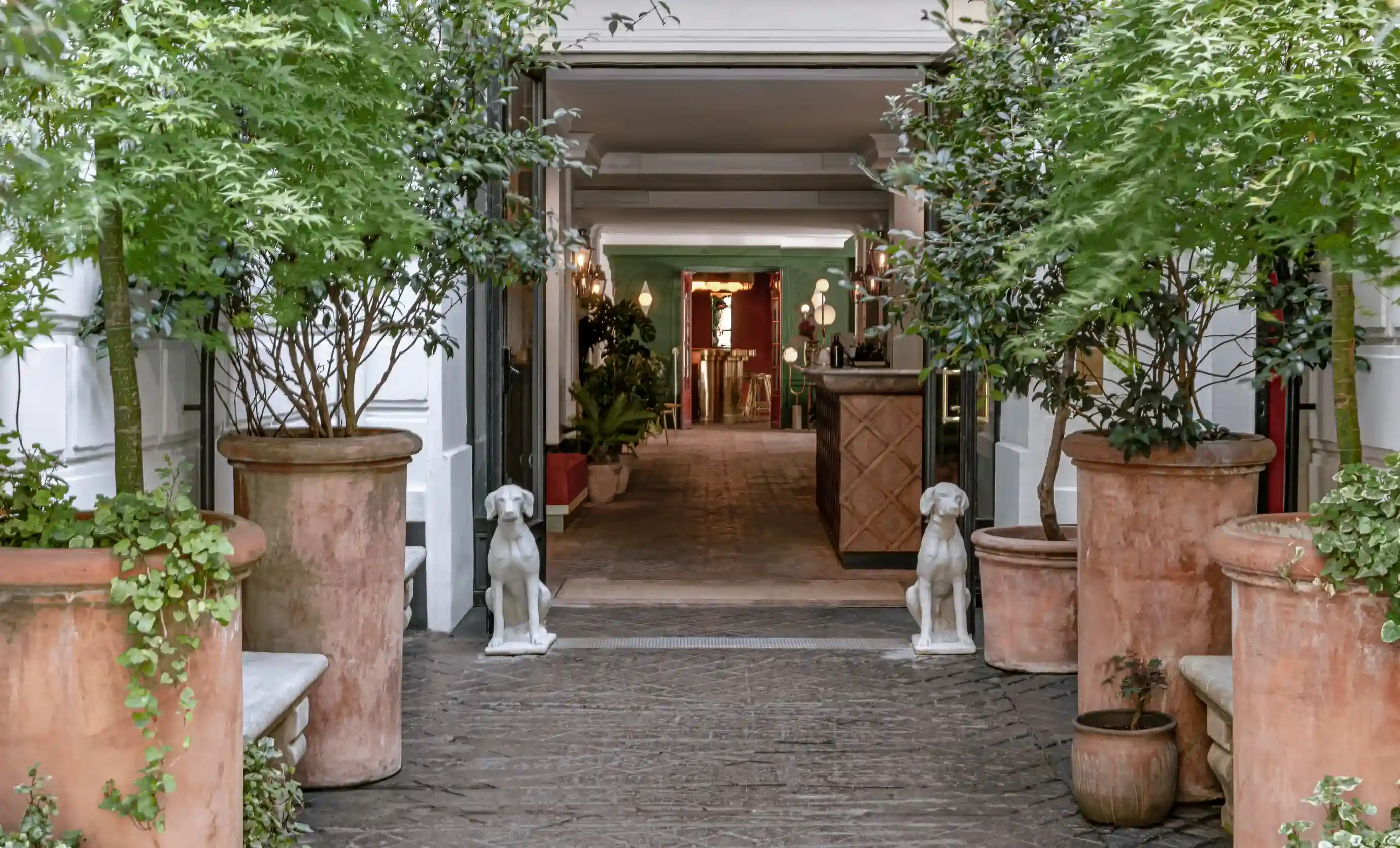 Entrada do hotel com vasos de plantas e duas estátuas de cães brancos flanqueando a porta.