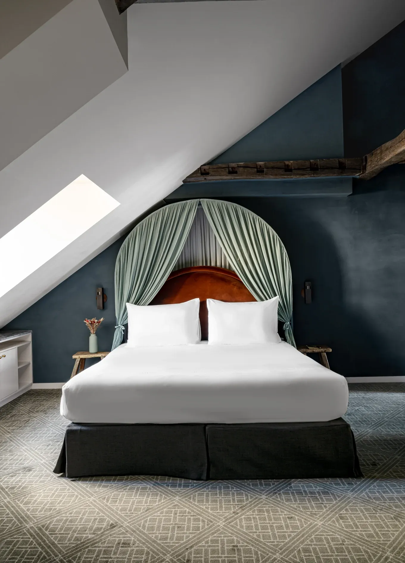 Cama com lençóis brancos, cabeceira marrom e cortinas verdes em um quarto de teto inclinado com paredes escuras.