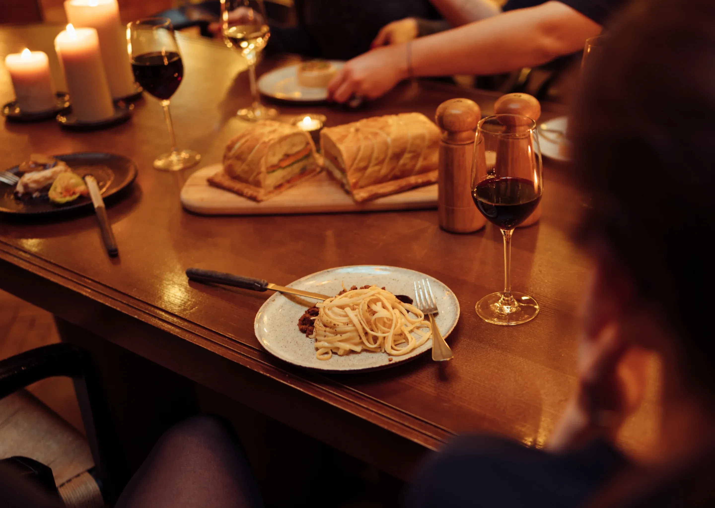 Mesa de jantar com macarrão, pão, taças de vinho tinto e branco, saleiros e pimenteiros e velas acesas.