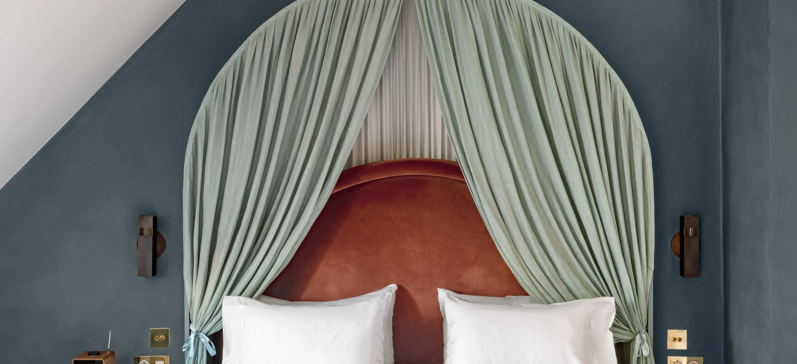 Cama com travesseiros brancos, cabeceira cor de ferrugem e cortinas verde-claras amarradas nas paredes azuis.
