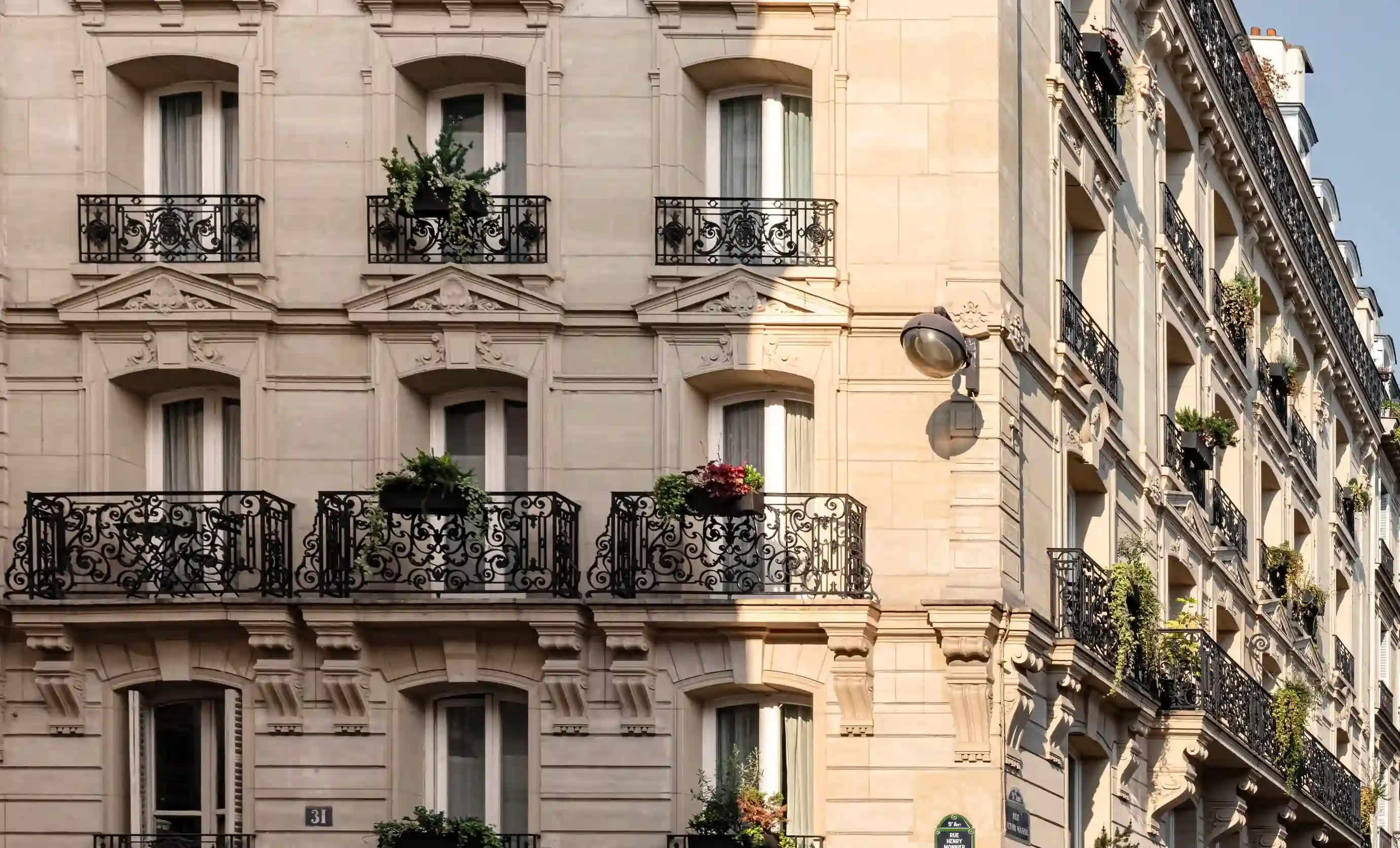 Edifício histórico de pedra bege com varandas de ferro ornamentadas e vasos de plantas em uma rua da cidade.