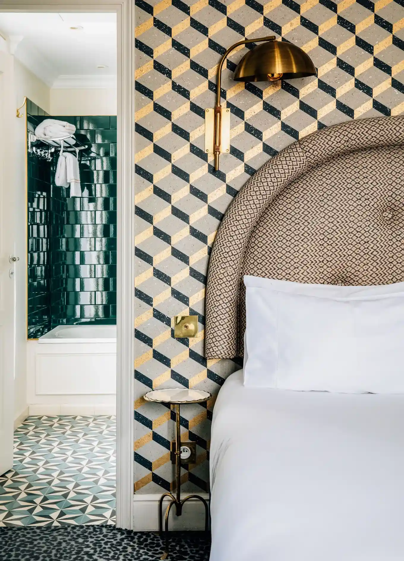 Canto do quarto de hotel com cabeceira estampada, roupa de cama branca, luminária de latão e vista para um banheiro de azulejos.