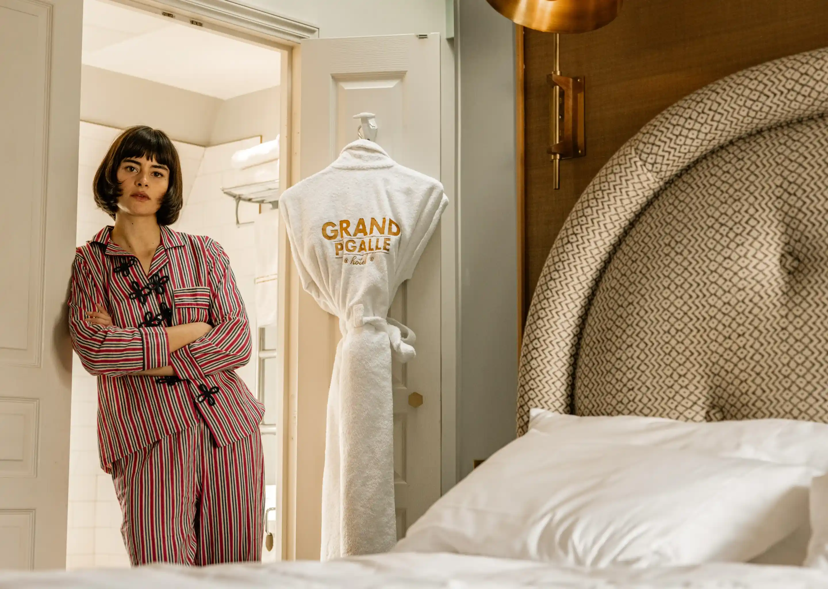 Mulher de pijama listrado encostada no batente de uma porta em um quarto de hotel com um roupão pendurado na porta.