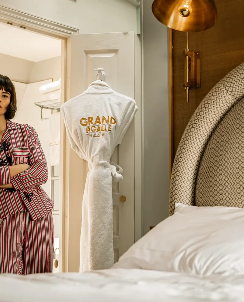Mulher de pijama listrado está perto de uma porta com uma túnica branca pendurada com o texto do Grand Pigalle Hotel.