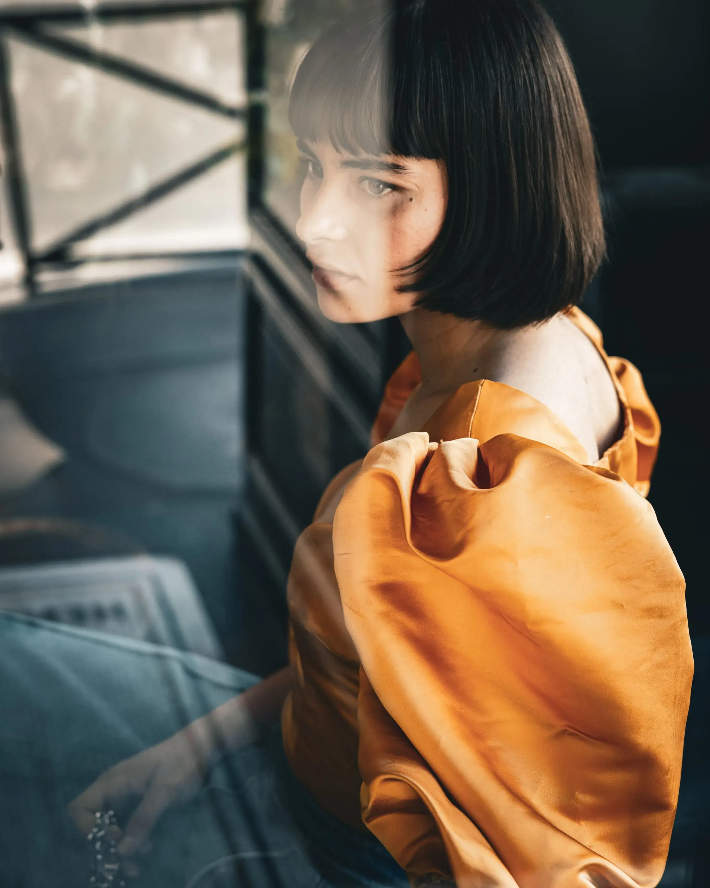 Mulher com cabelo curto e escuro e blusa laranja de manga bufante vista através de uma janela refletiva.