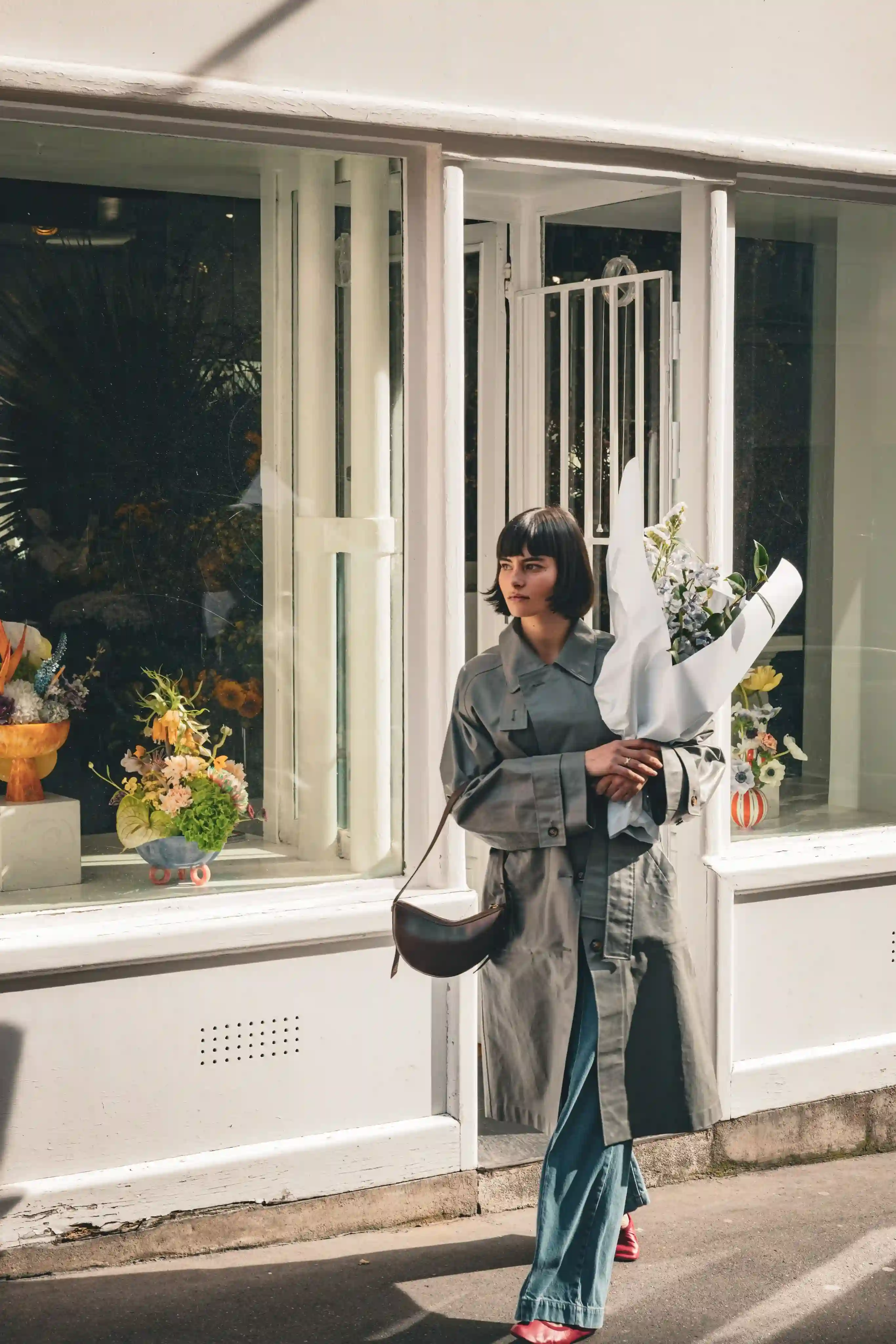 Mulher com um sobretudo cinza segurando um buquê passa pela vitrine de uma floricultura com arranjos florais.