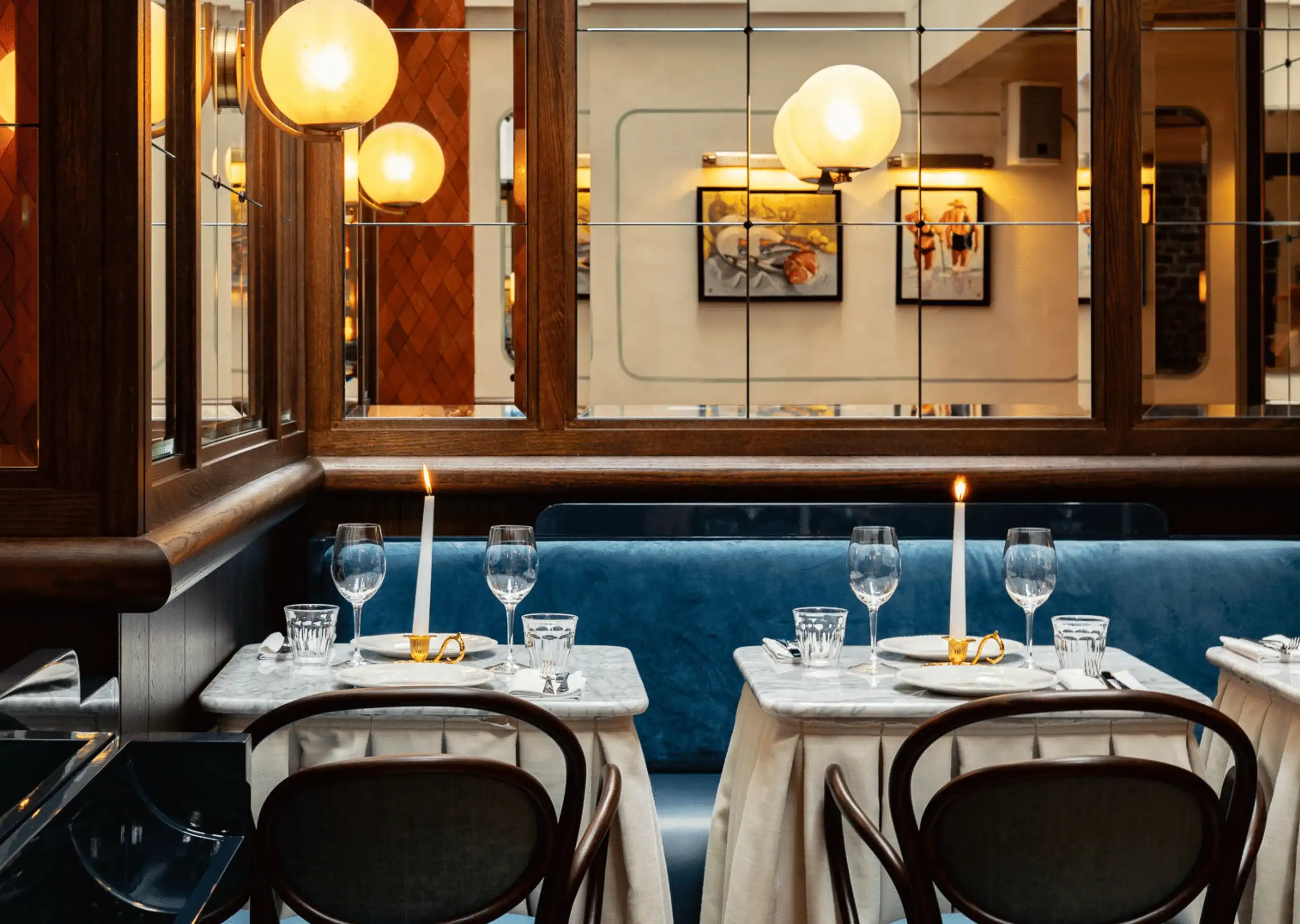 Comodi tavoli da ristorante con panca in velluto blu, lenzuola bianche, candele accese e parete a specchio con luci a globo.
