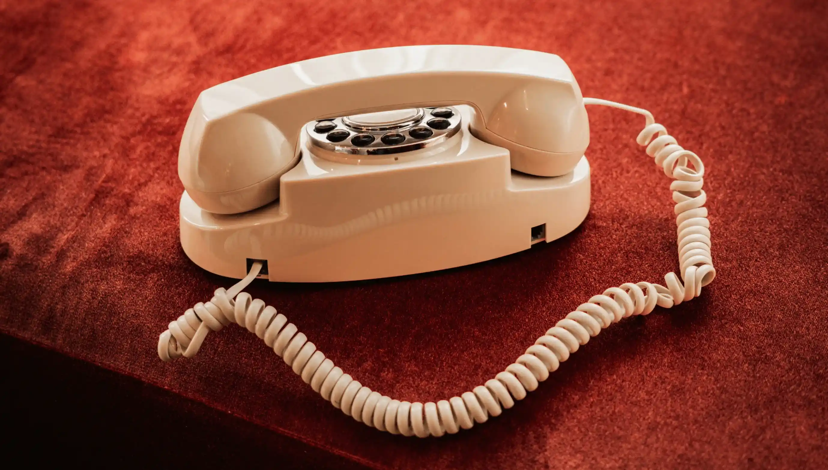 Telefono con linea rotativa beige con cavo a spirale appoggiato su una superficie di velluto rosso.