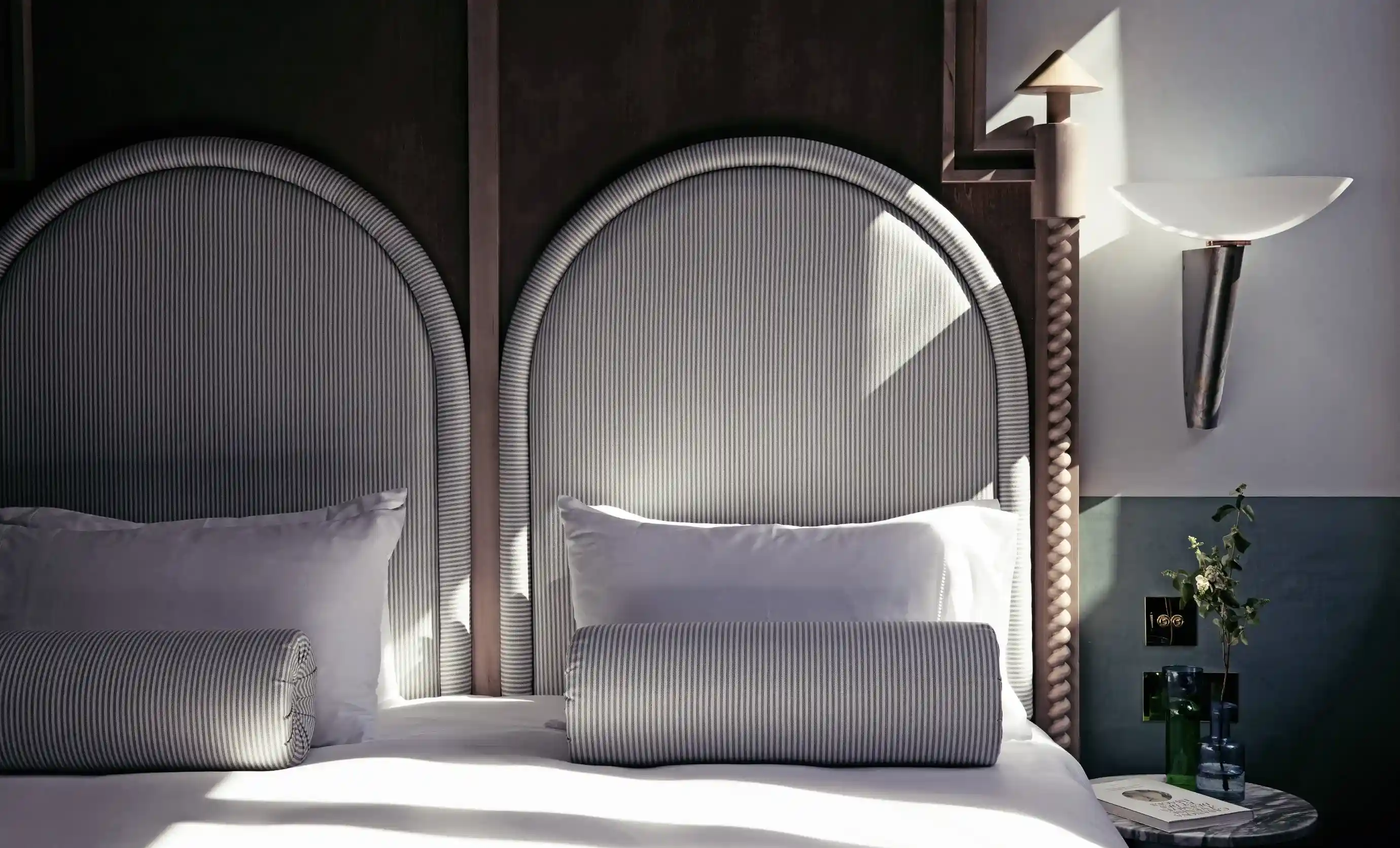 Cama de hotel iluminada pelo sol com cabeceiras listradas em arco, travesseiros brancos, almofadas, mesa lateral e arandela de parede.