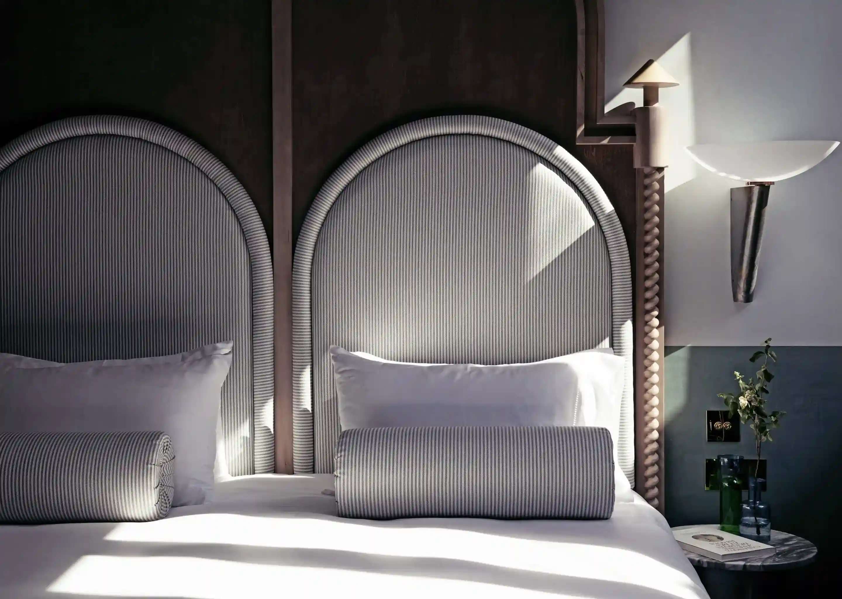 Quarto de hotel com duas camas individuais com cabeceiras listradas arredondadas, travesseiros e uma mesa de cabeceira com decoração.
