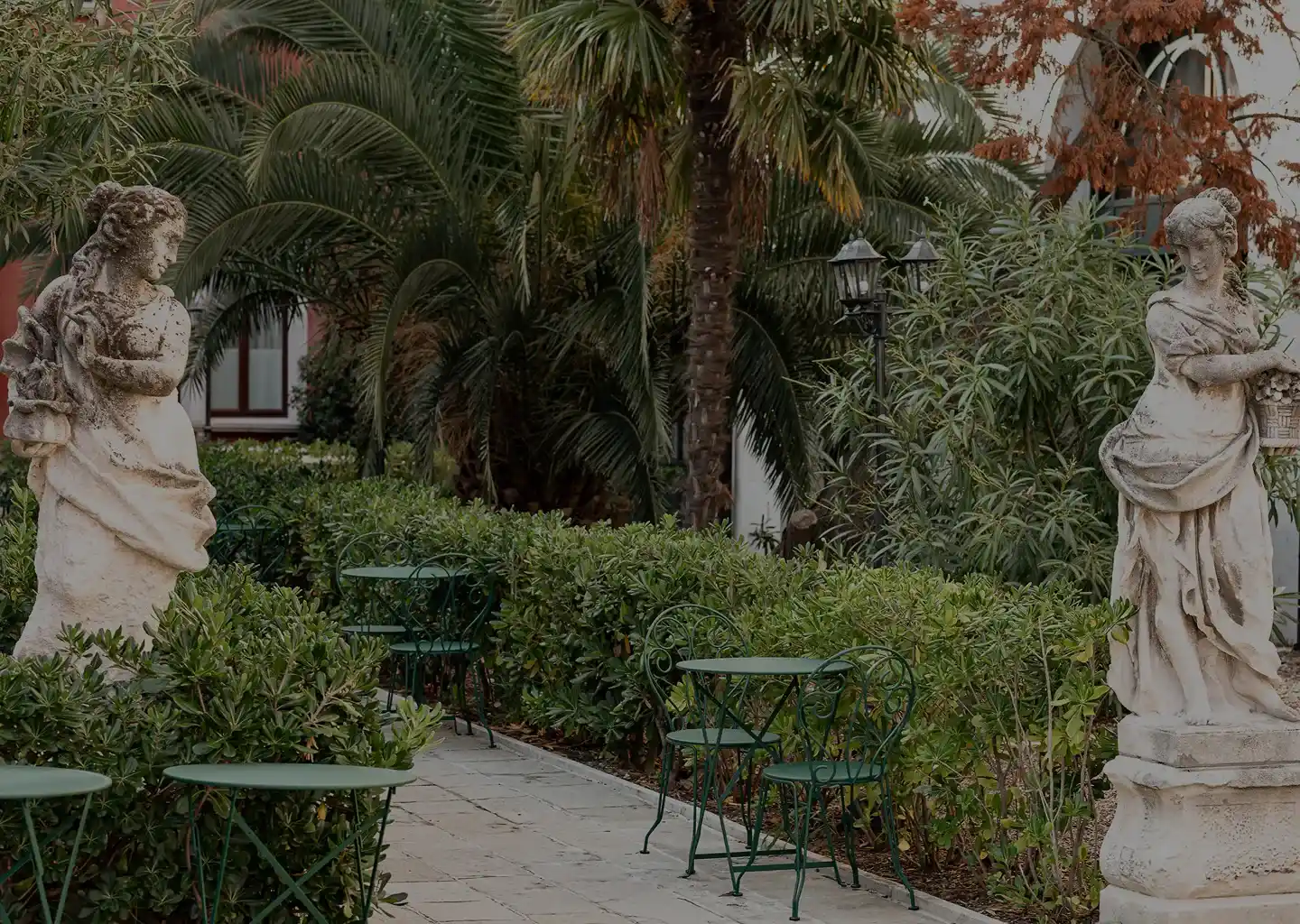 Mesas de jardim ao ar livre com mesas de metal verde, cadeiras, duas estátuas de pedra clássicas e vegetação exuberante.