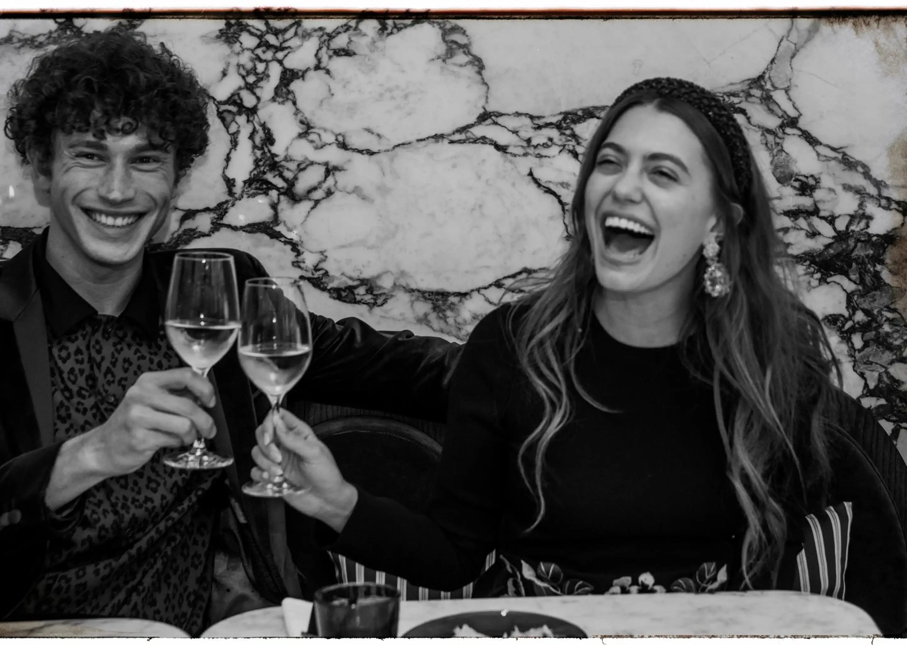 Homem e mulher sorridentes tilintando taças de vinho em um restaurante com uma parede de mármore atrás delas.