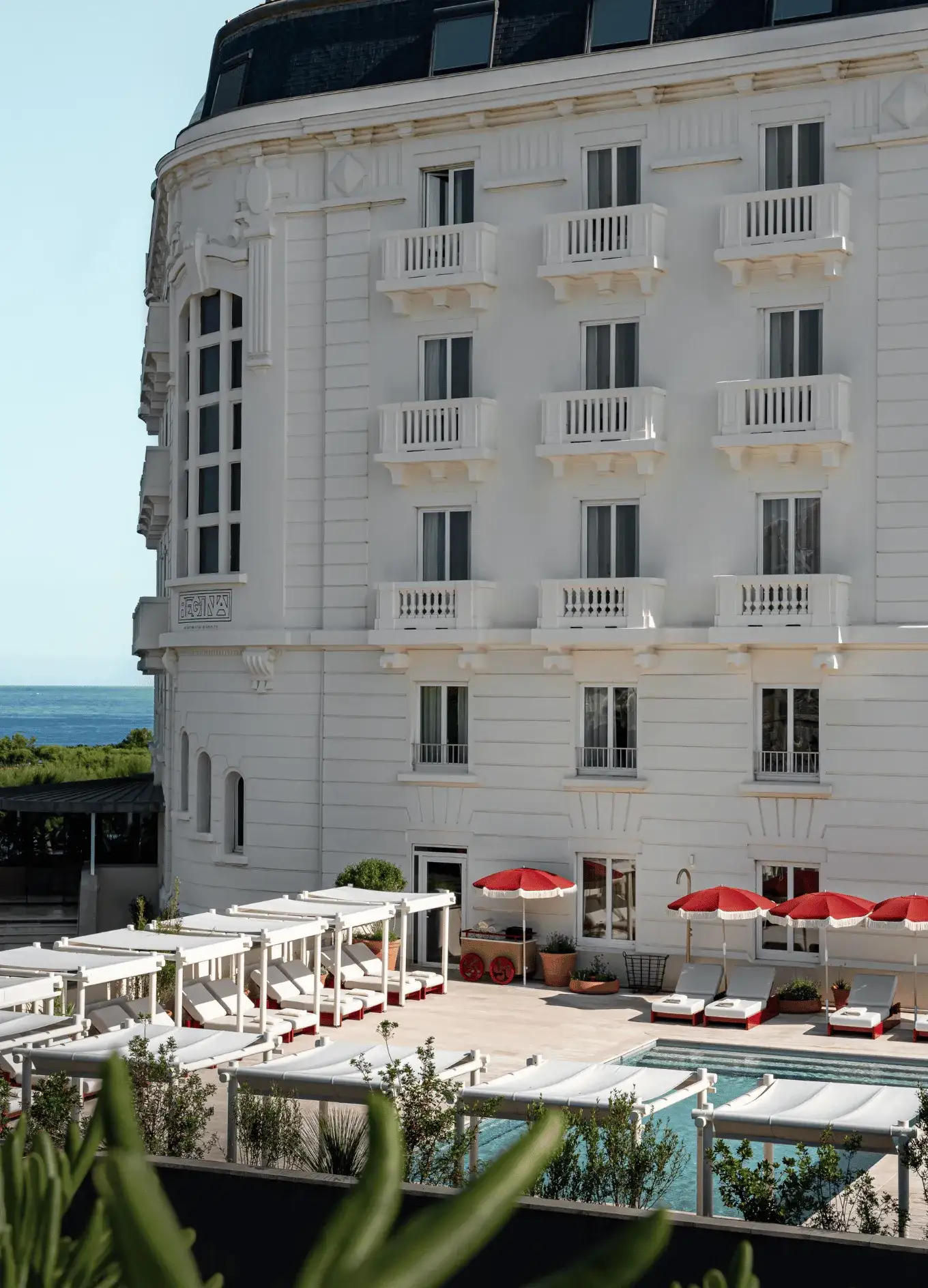 Edificio alberghiero bianco con balconi accanto a una piscina con lettini e ombrelloni rossi in riva al mare.