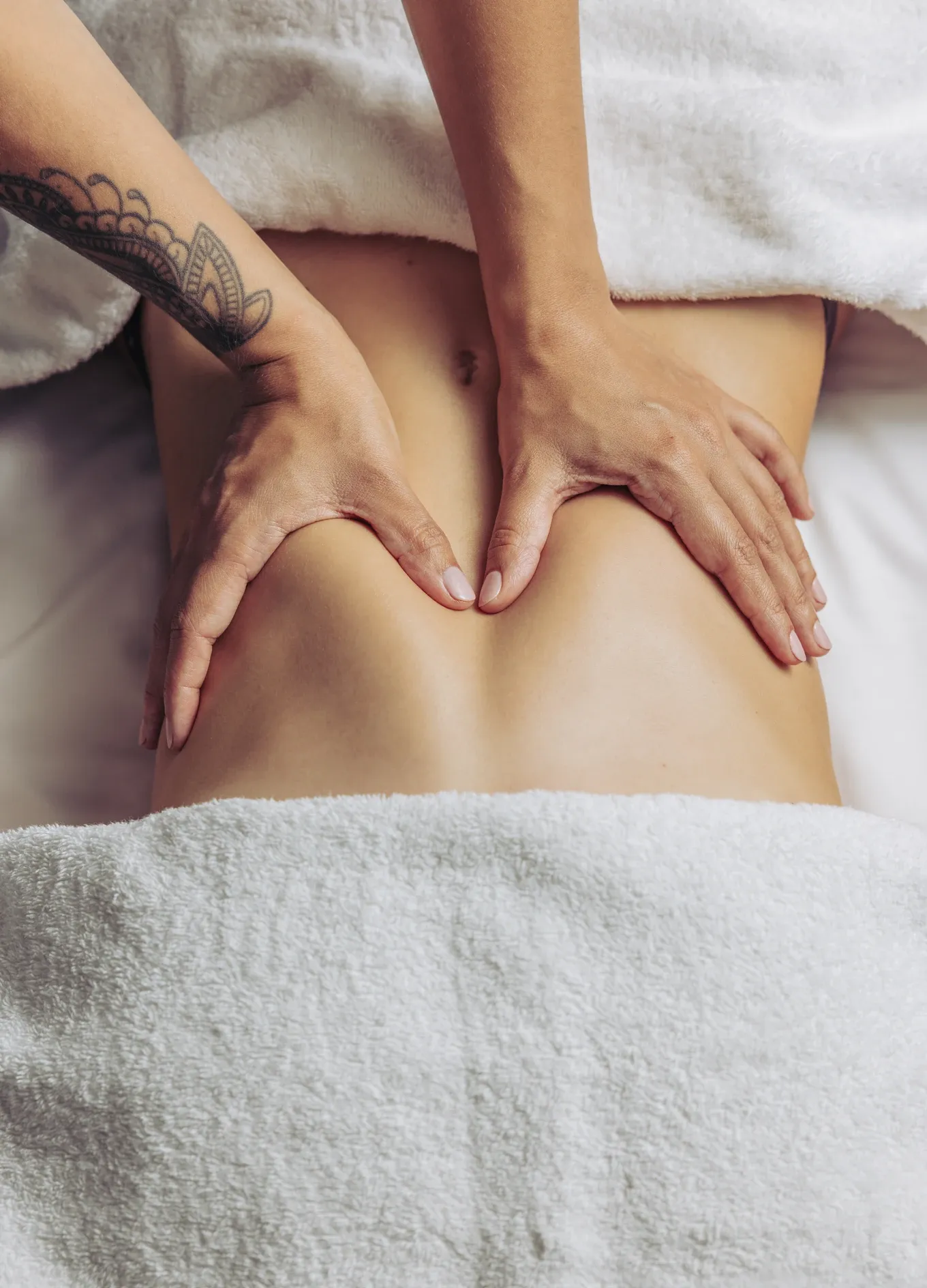 Mani con tatuaggio che massaggiano la parte bassa della schiena nuda di una persona coperta parzialmente con asciugamani bianchi.