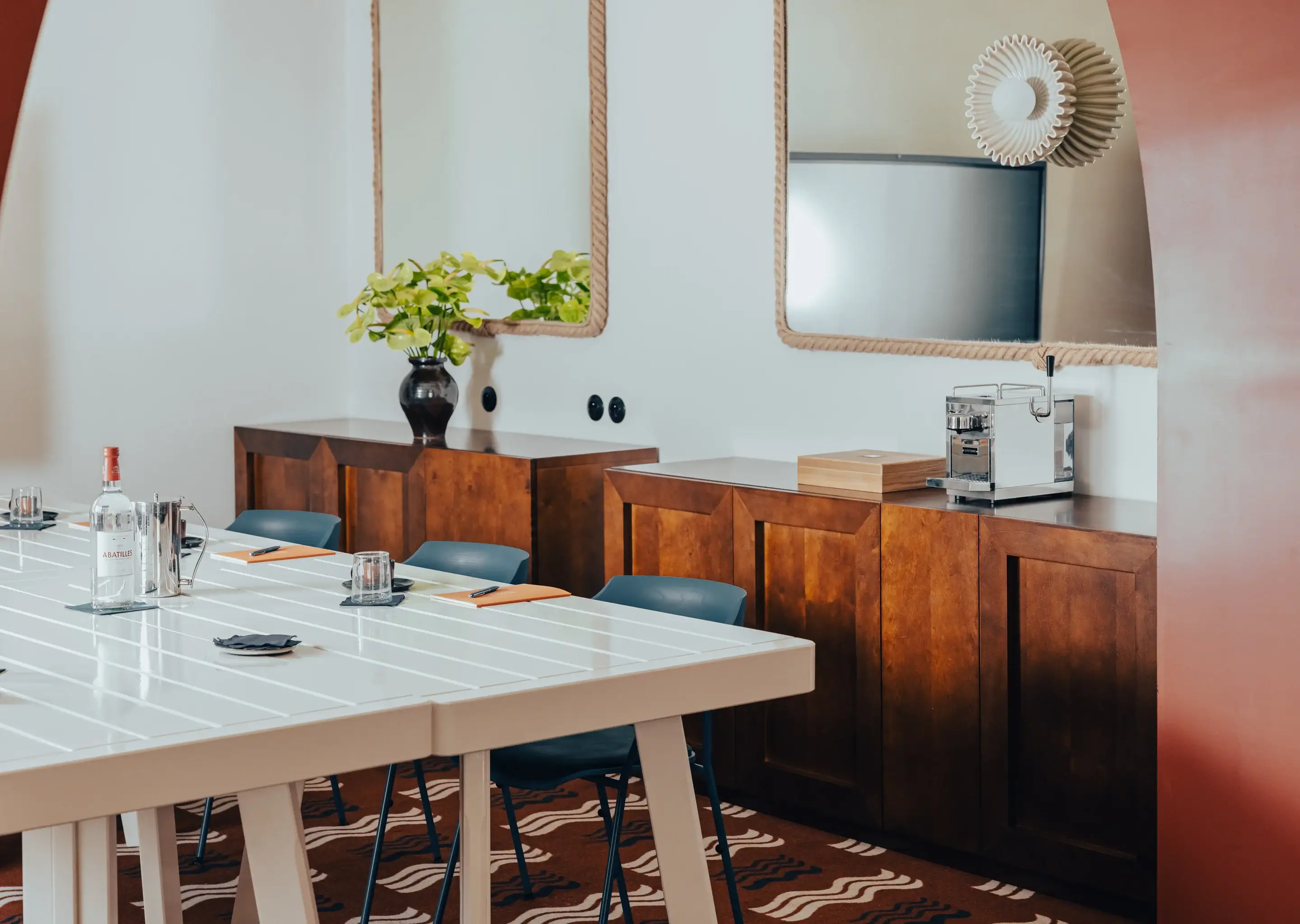 Moderna sala riunioni con tavolo bianco, sedie blu, armadi in legno, pianta verde e macchina da caffè.